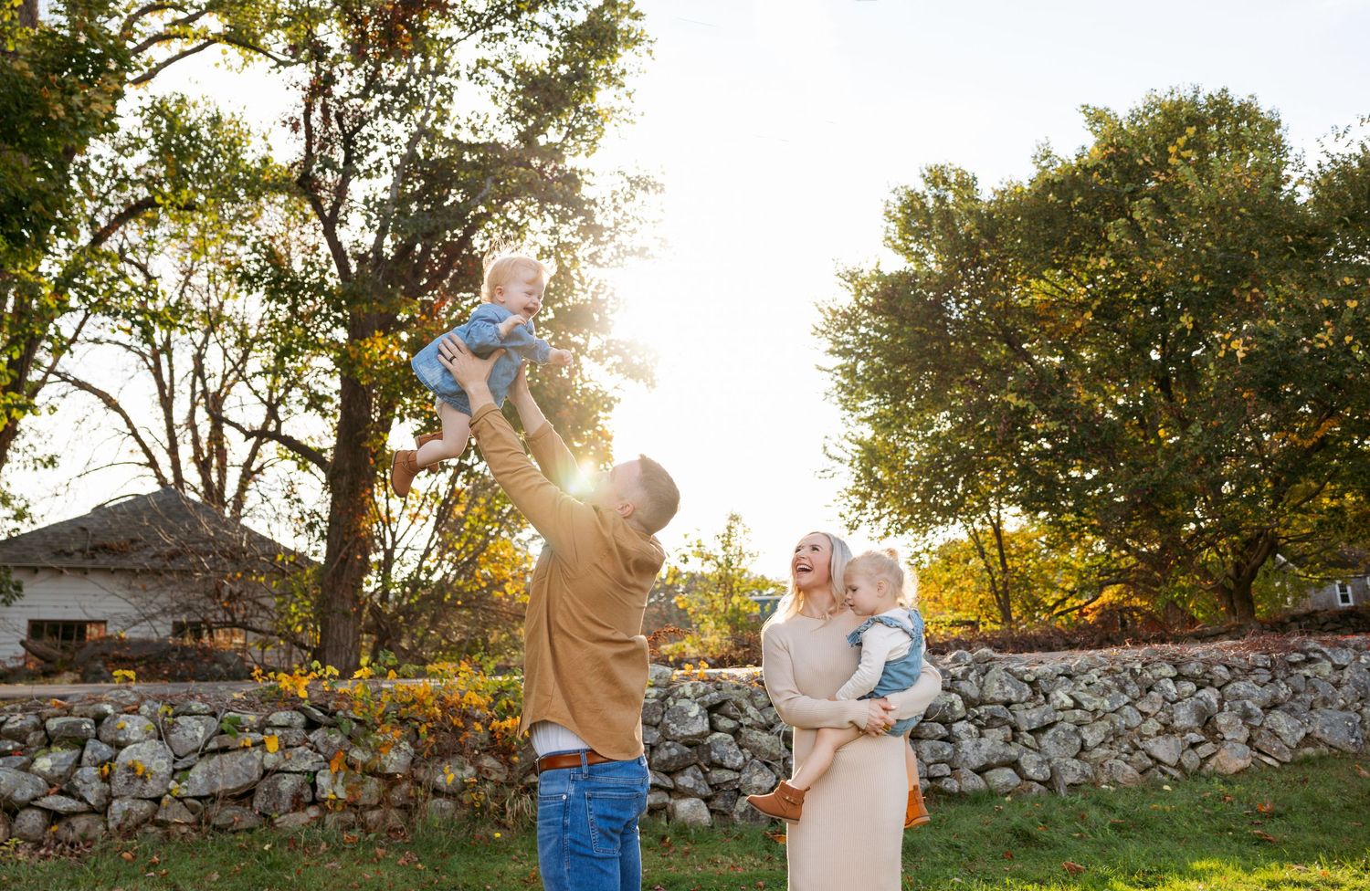 A couple walks together against a sunset backdrop with trees and a stone wall during an autumn evening photo session.