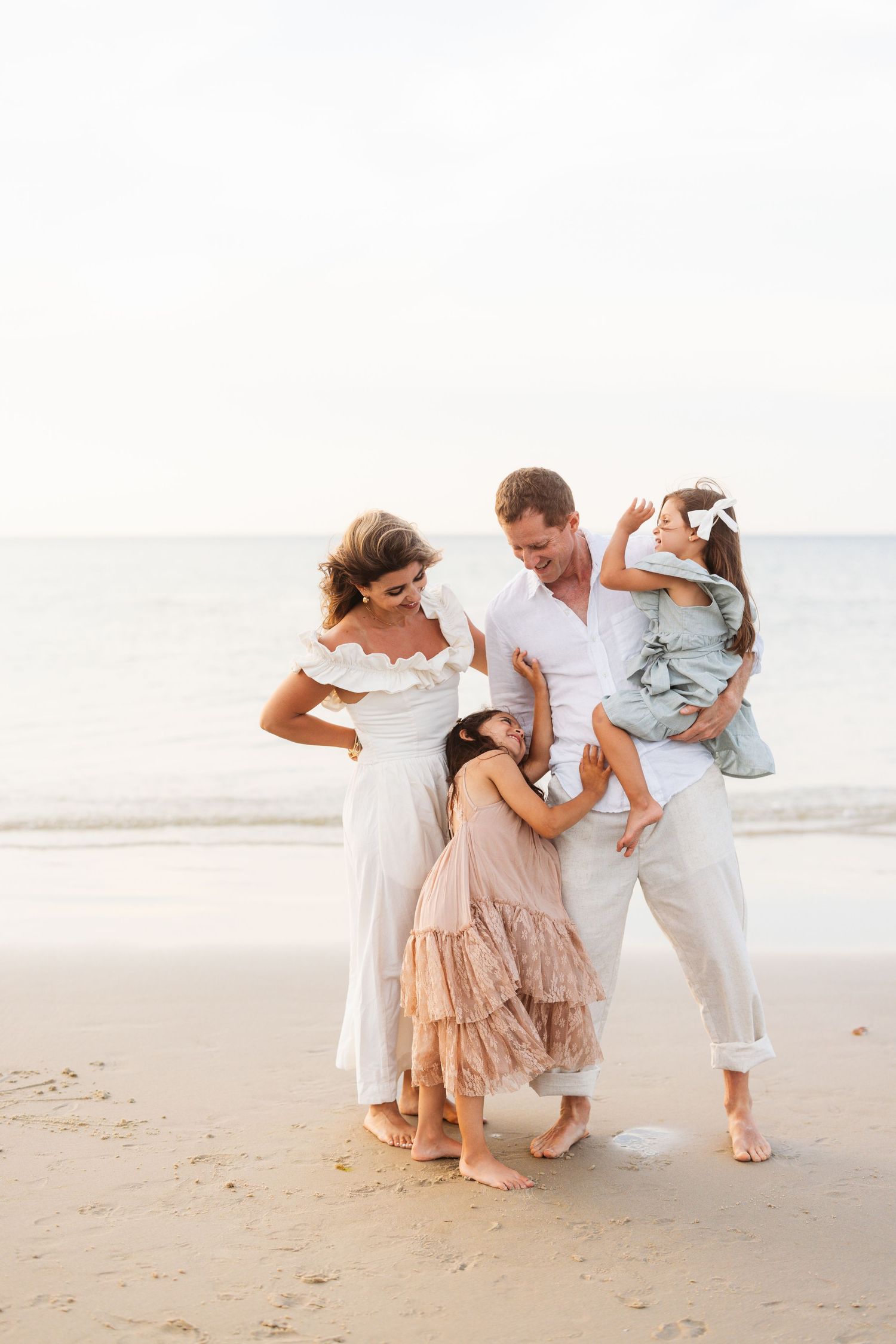 Family playing and dancing together on a sunny beach in coordinated white and blush outfits.