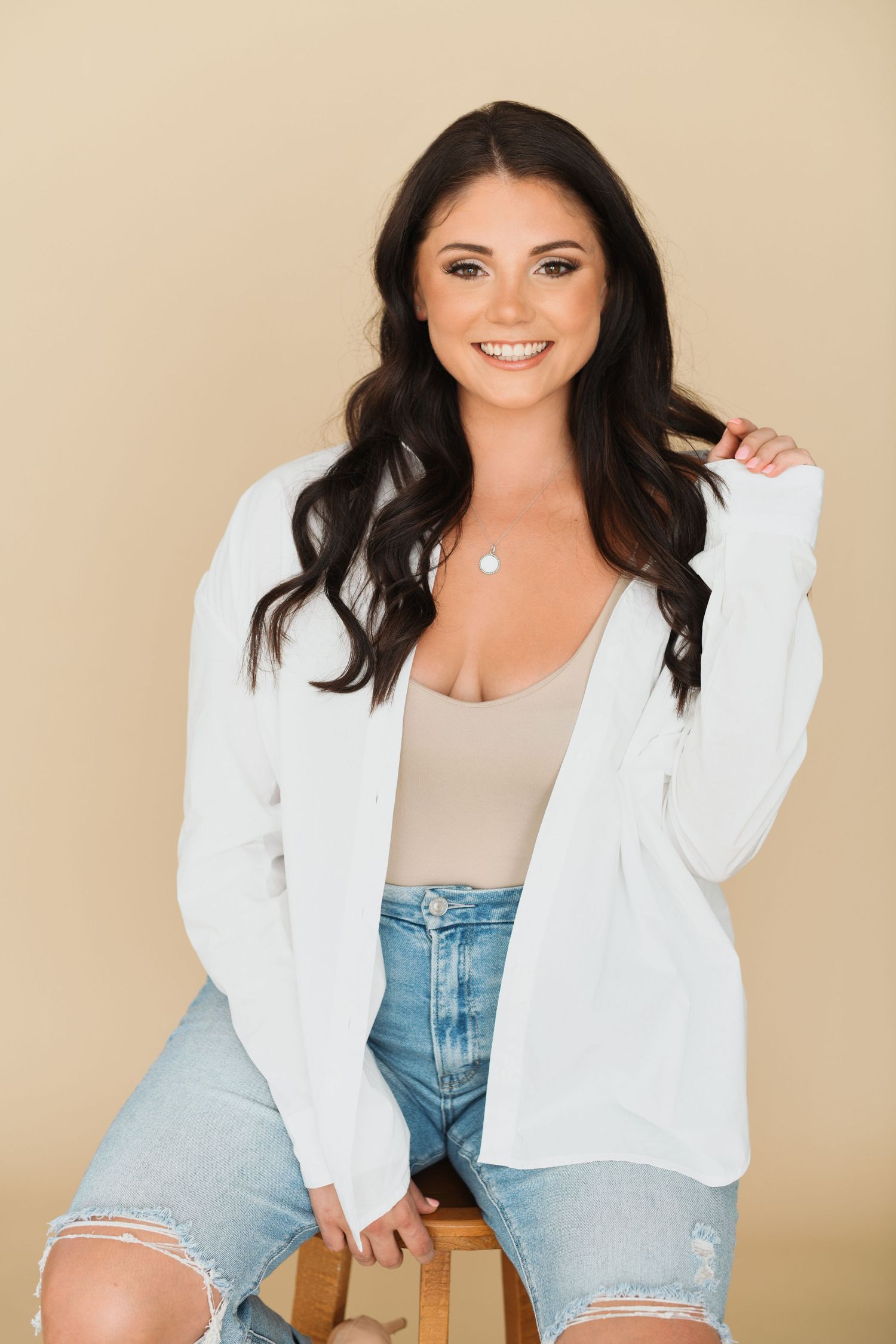 Smiling brunette woman in white cardigan, beige top, and ripped jeans sitting on stool against tan background.