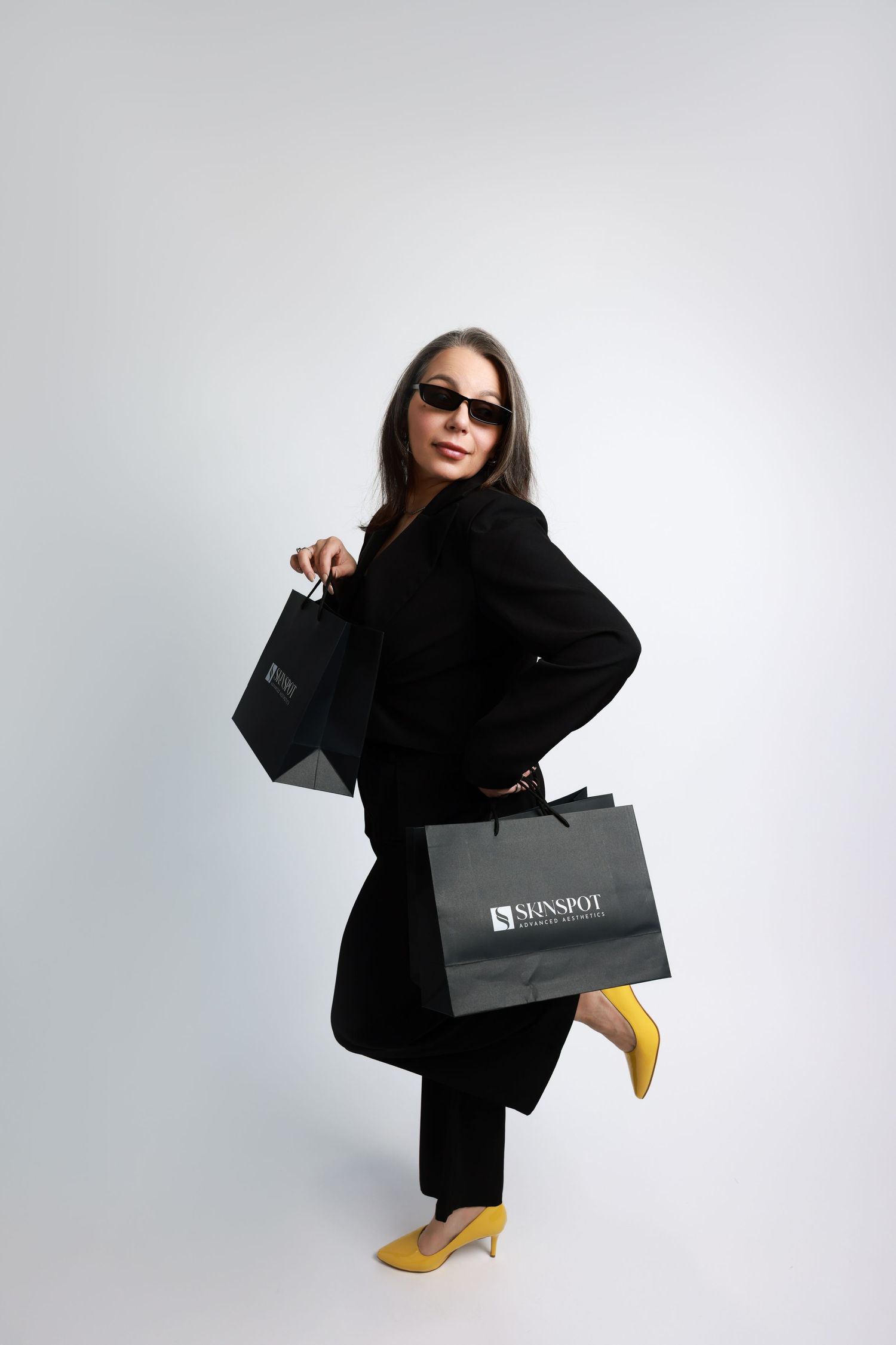 Stylish person in black outfit holds designer shopping bags while posing against white background.