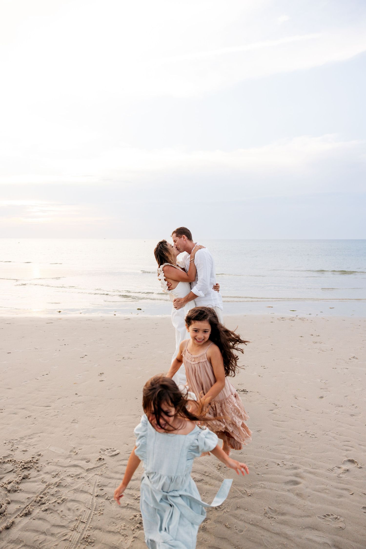 People in light flowy outfits dance and play together on a sandy beach at sunset.
