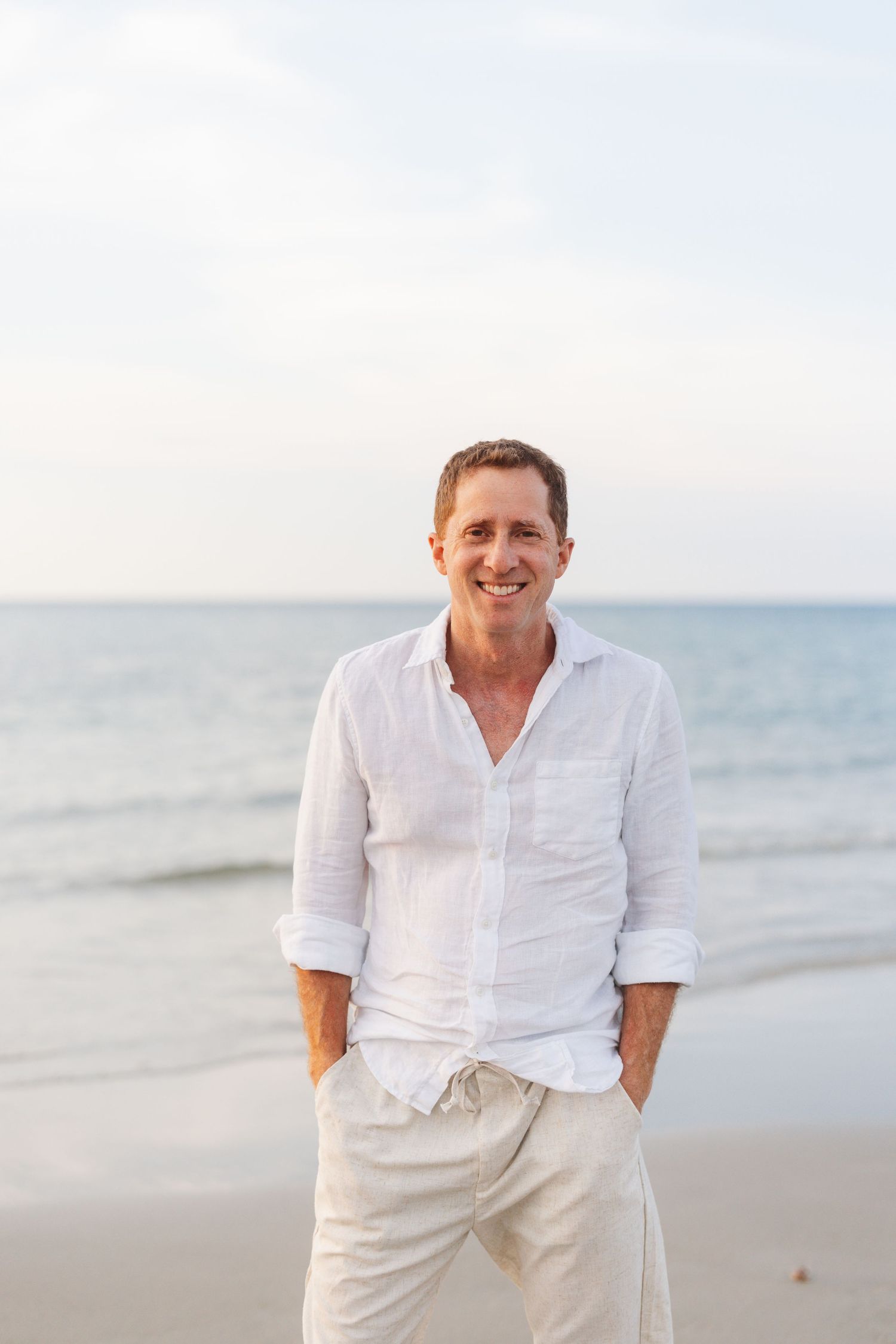 A person in white attire posing casually on a serene beach with ocean waves in the background.