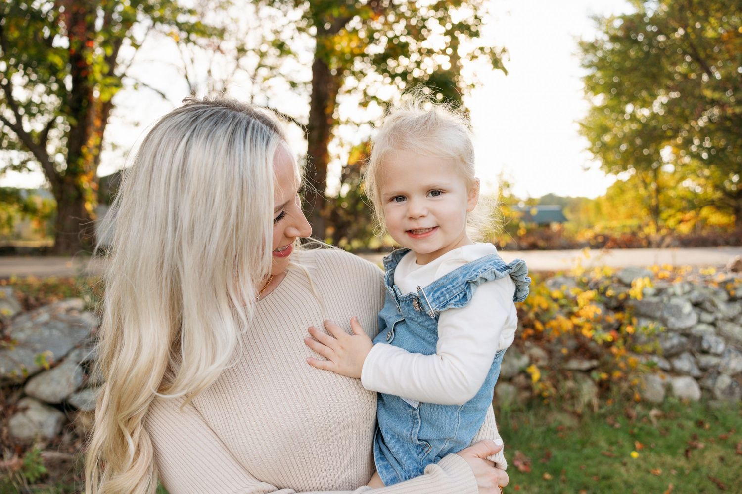 Loving embrace captured during outdoor autumn photoshoot with rustic stone wall and trees in background.