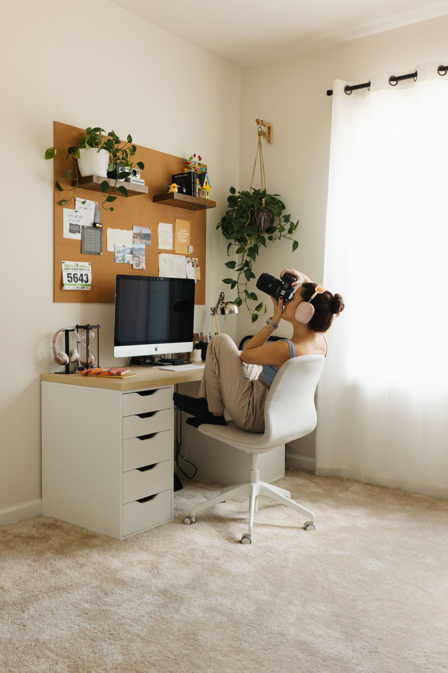 Modern home office setup with white desk, wooden shelving, plants and plush beige carpeting along wall in natural light.