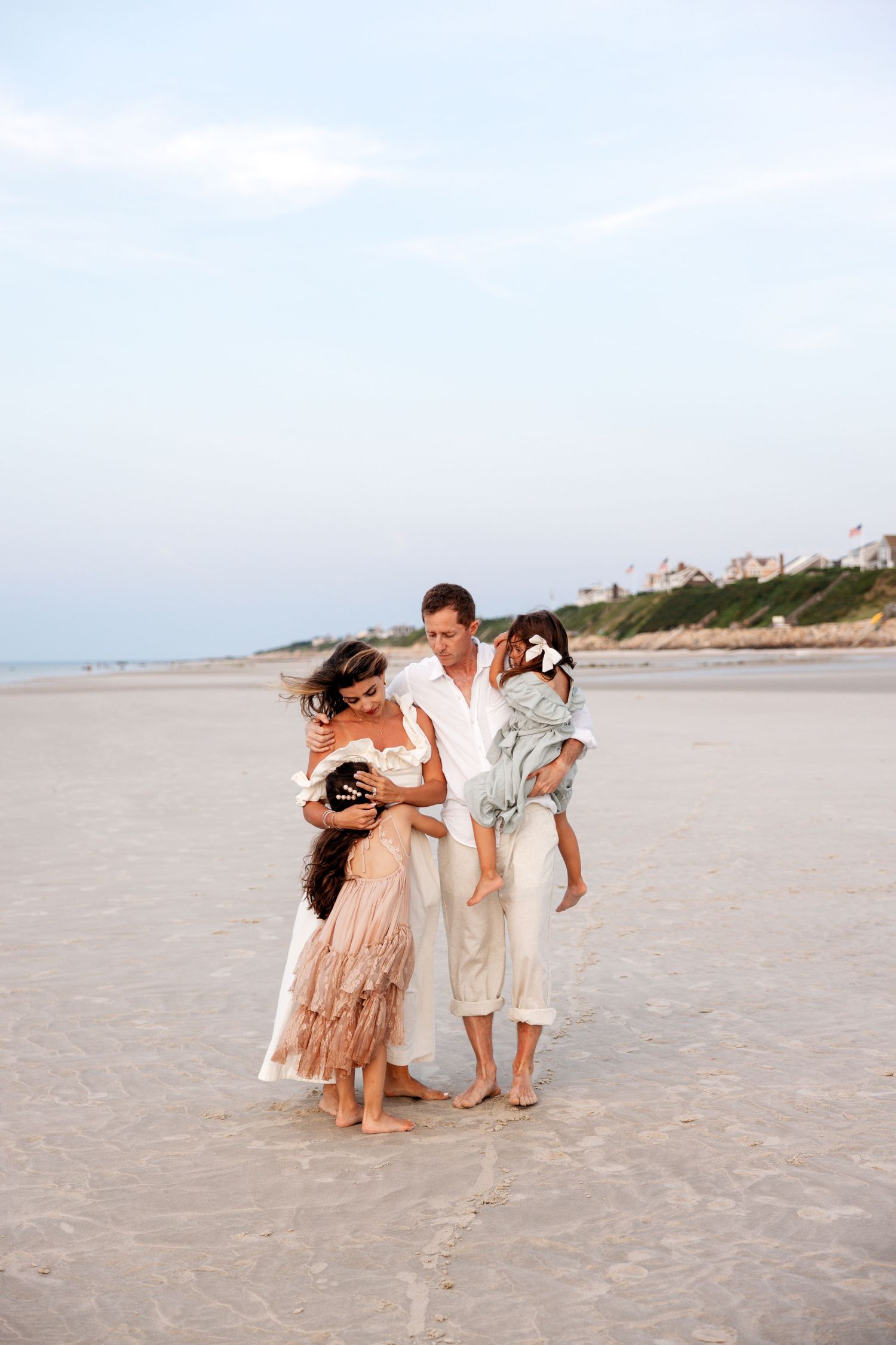 Family in coordinated beige and pink outfits sharing playful moments together on a sandy beach at sunset.