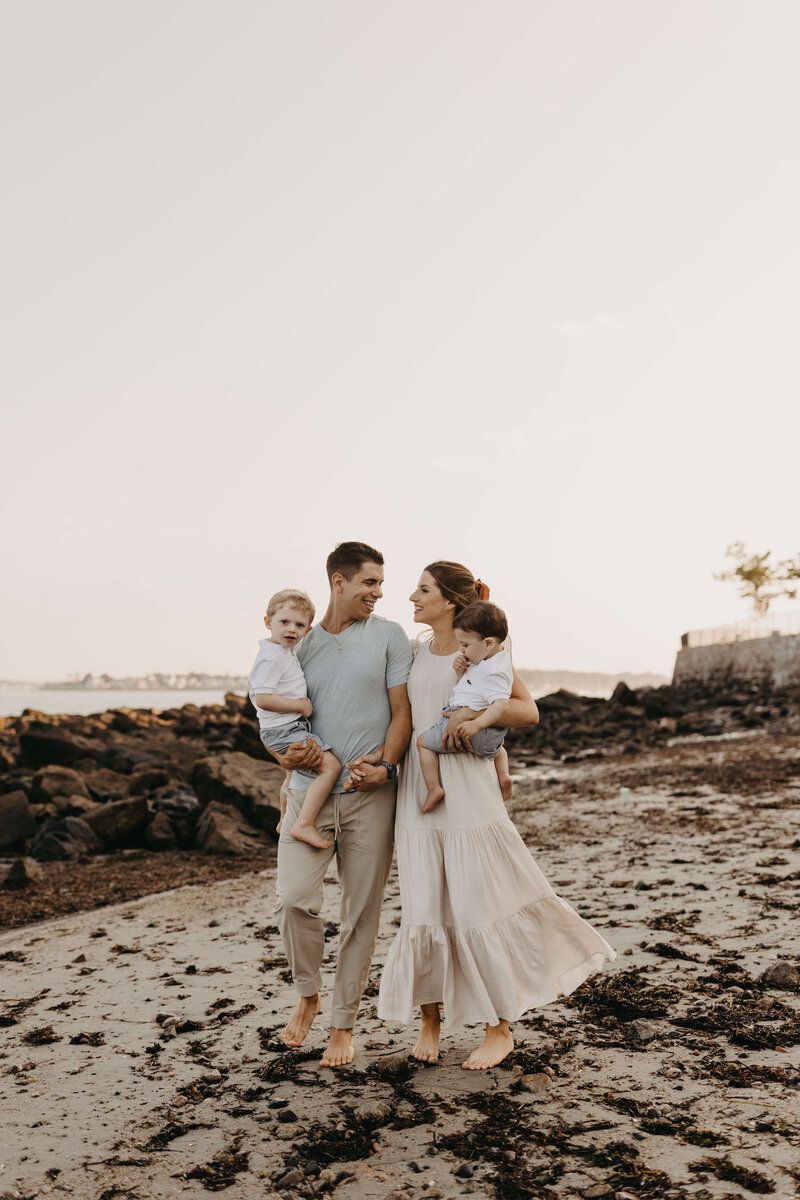 Family in coordinating neutral outfits walking together on rocky beach at sunset.