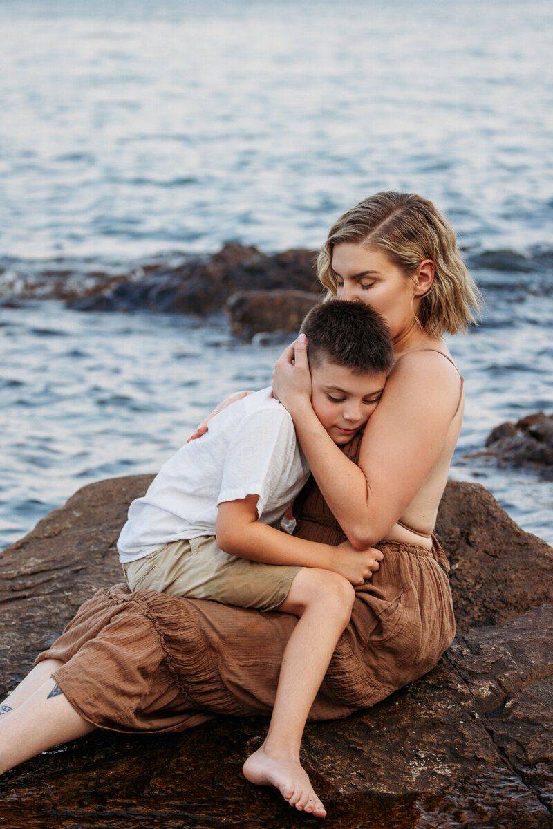 Intimate seaside photoshoot with two people cuddling on rocky beach at sunset.