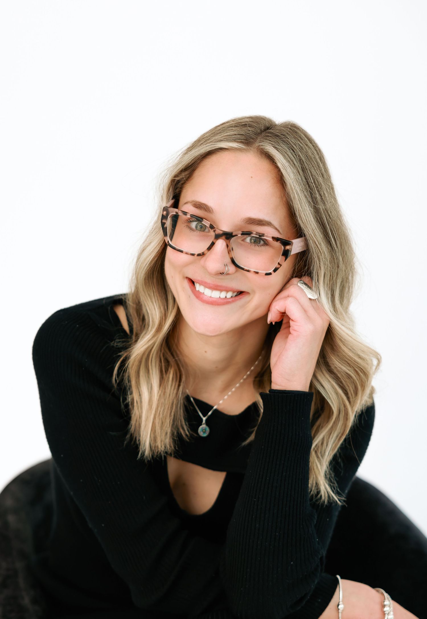 Smiling blonde woman with tortoiseshell glasses in black top posing against white background.