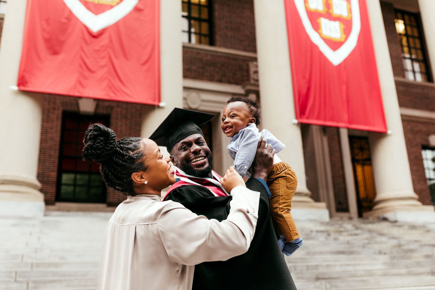 Festive celebration with people hugging in front of Harvard University's iconic red banners.