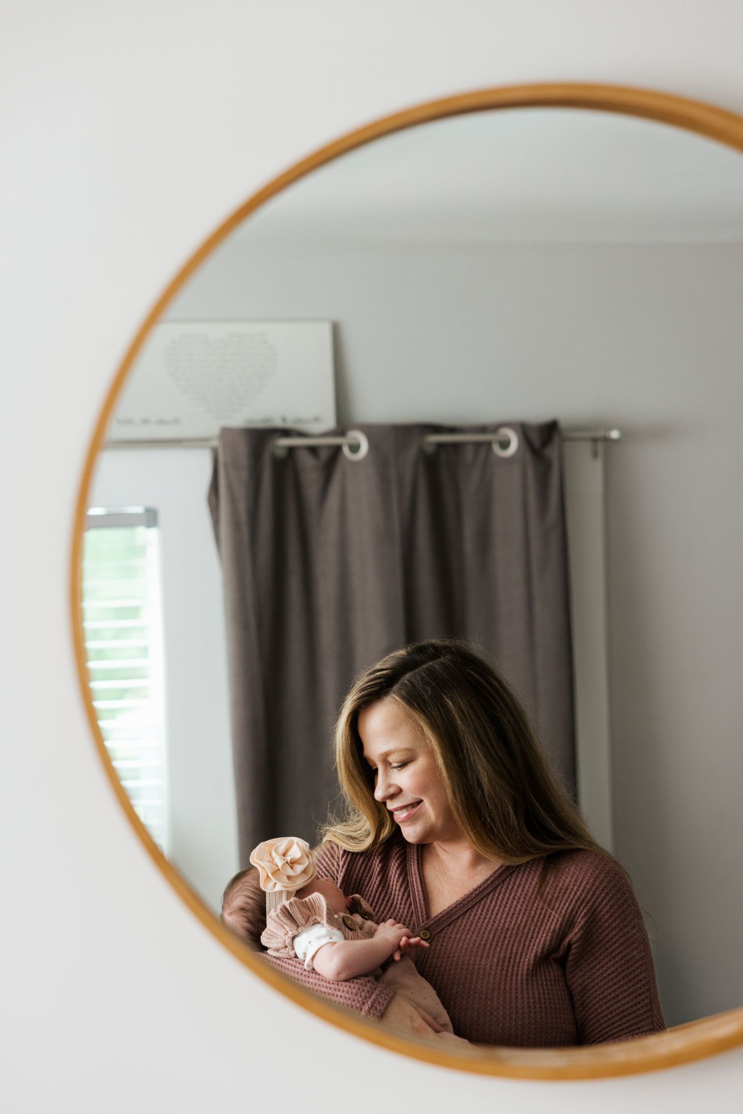 Three oval mirrors on a wall reflect a person in a brown sweater sitting beside curtained windows and a wall mirror collection.