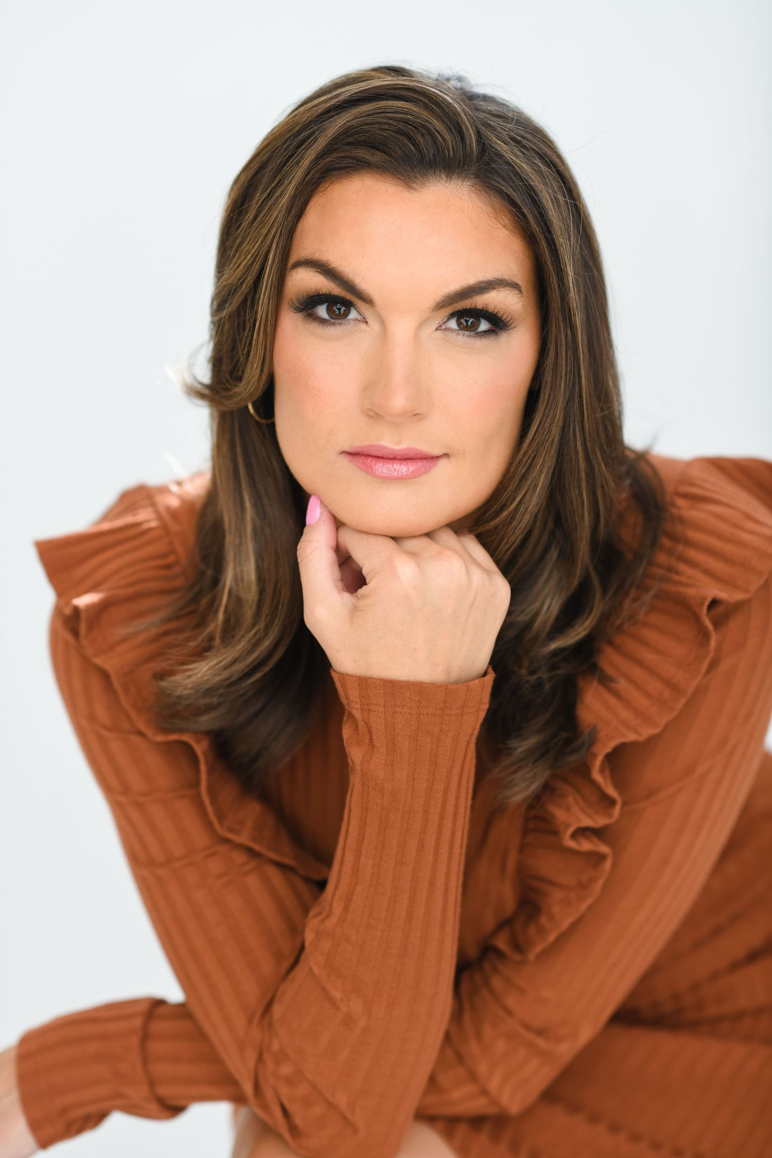 Brunette woman in rust ruffled sweater resting chin on hand with smoky eye makeup in studio.