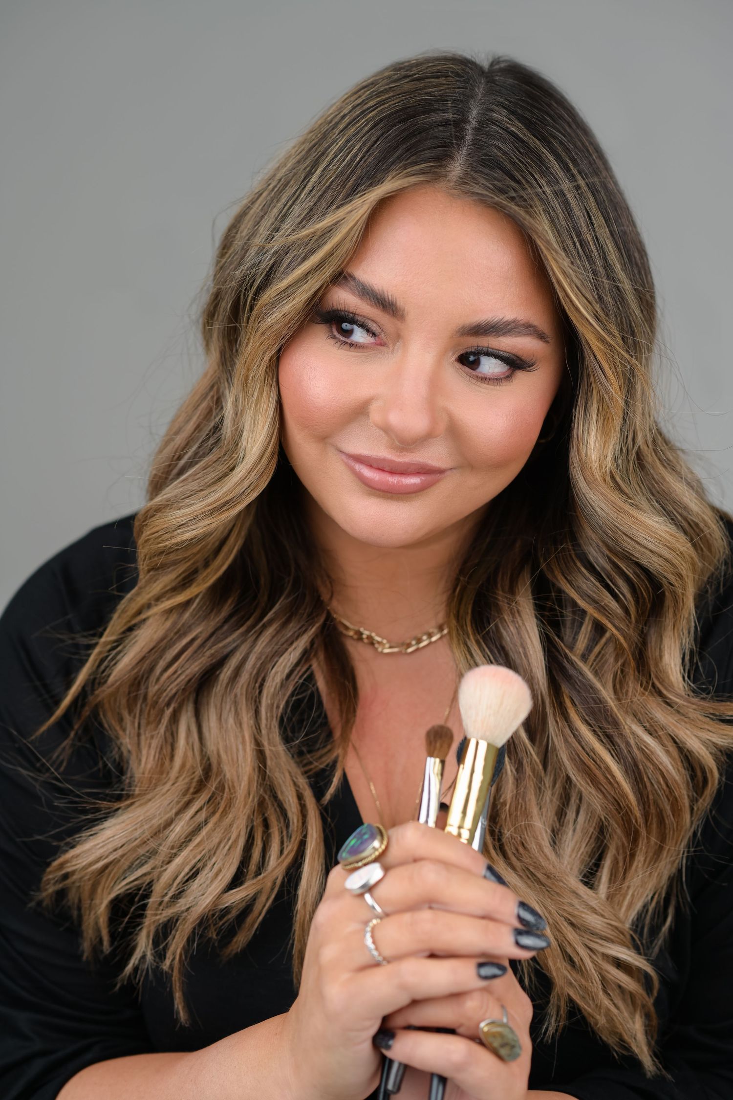 Woman with wavy balayage hair holding makeup brushes, wearing black top and gold necklace, gray background.