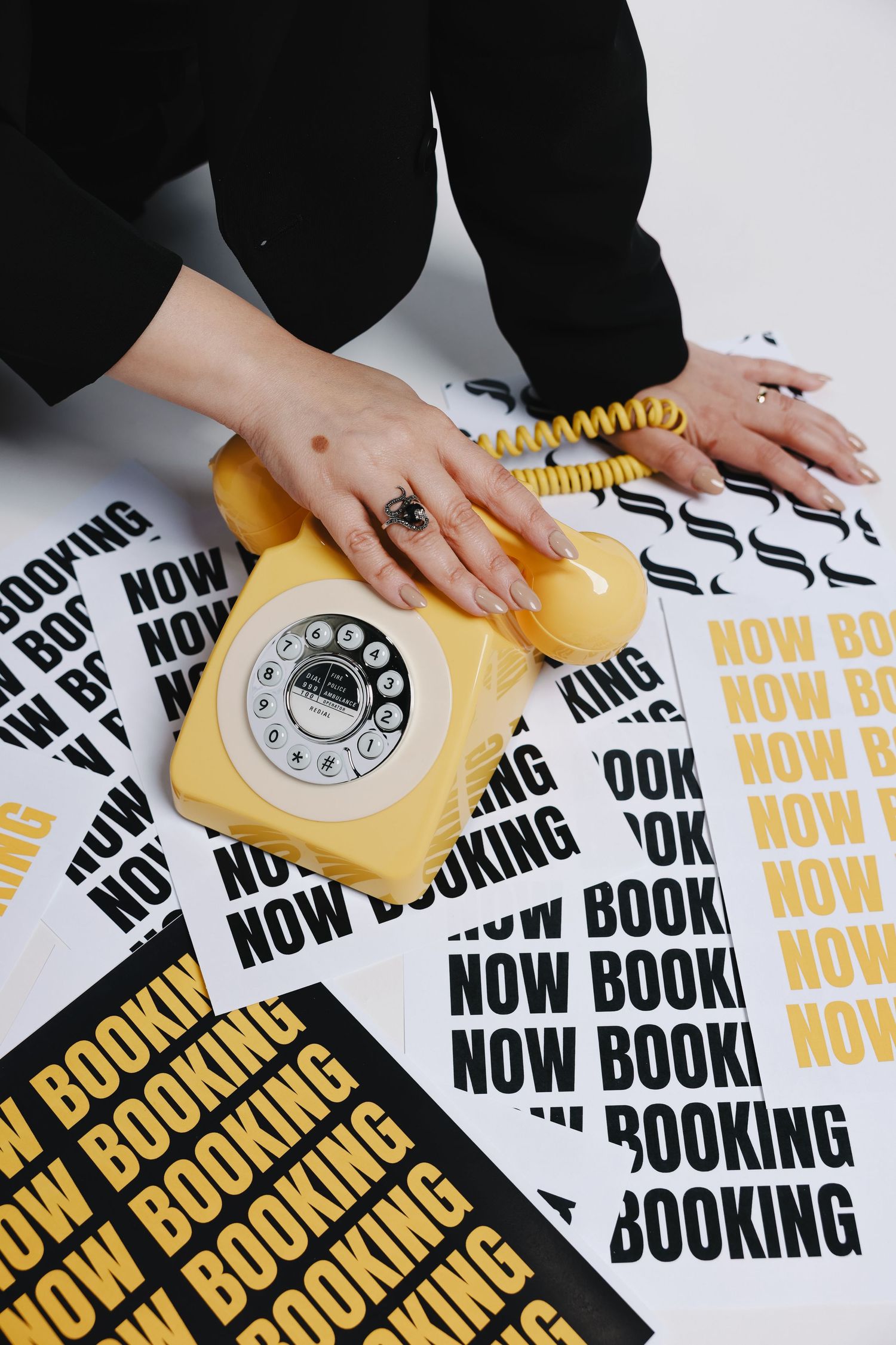 Vintage yellow rotary telephone on papers with repetitive black and yellow 'NOW BOOKING' text design.