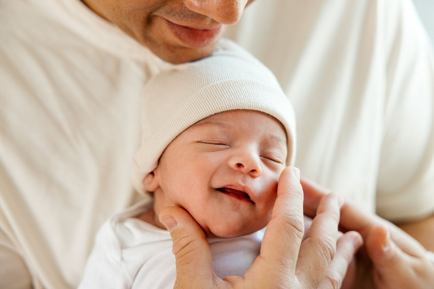 A newborn baby wearing a white hat smiles while being held.