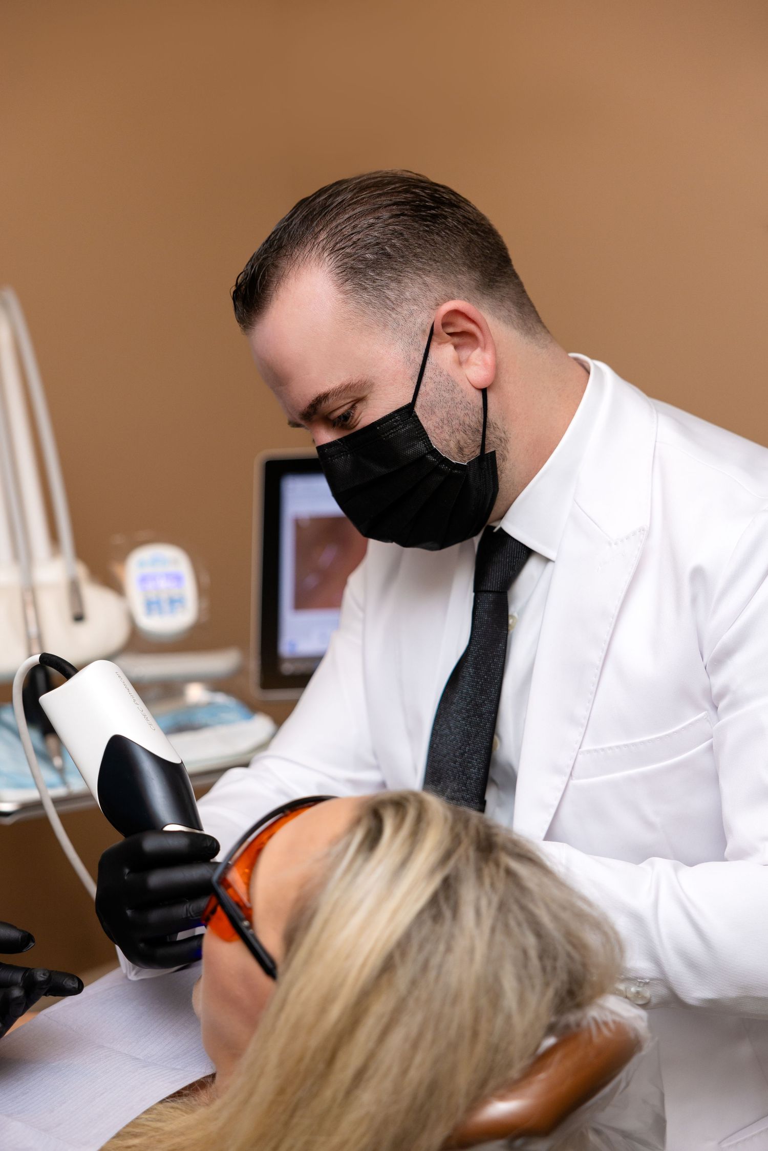 A dermatologist in a white coat and black mask performs laser skin treatment on a female patient wearing protective eyewear.