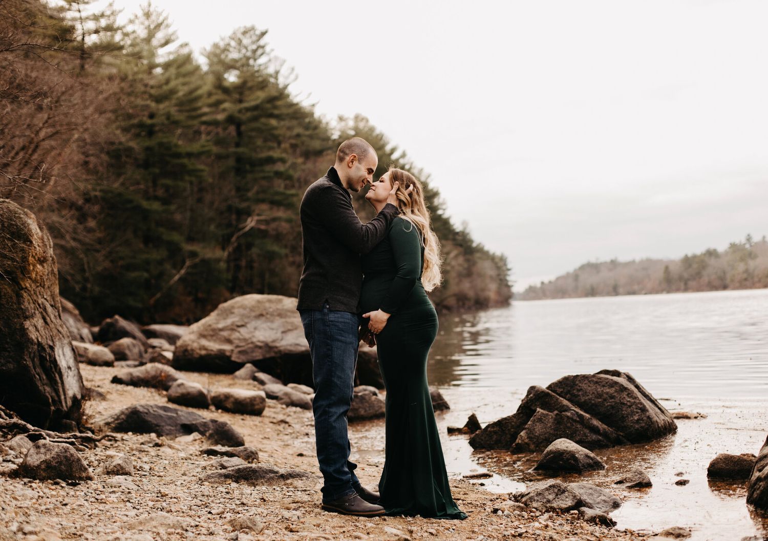Couple sharing tender moments along a rocky waterfront with pine trees in the background.