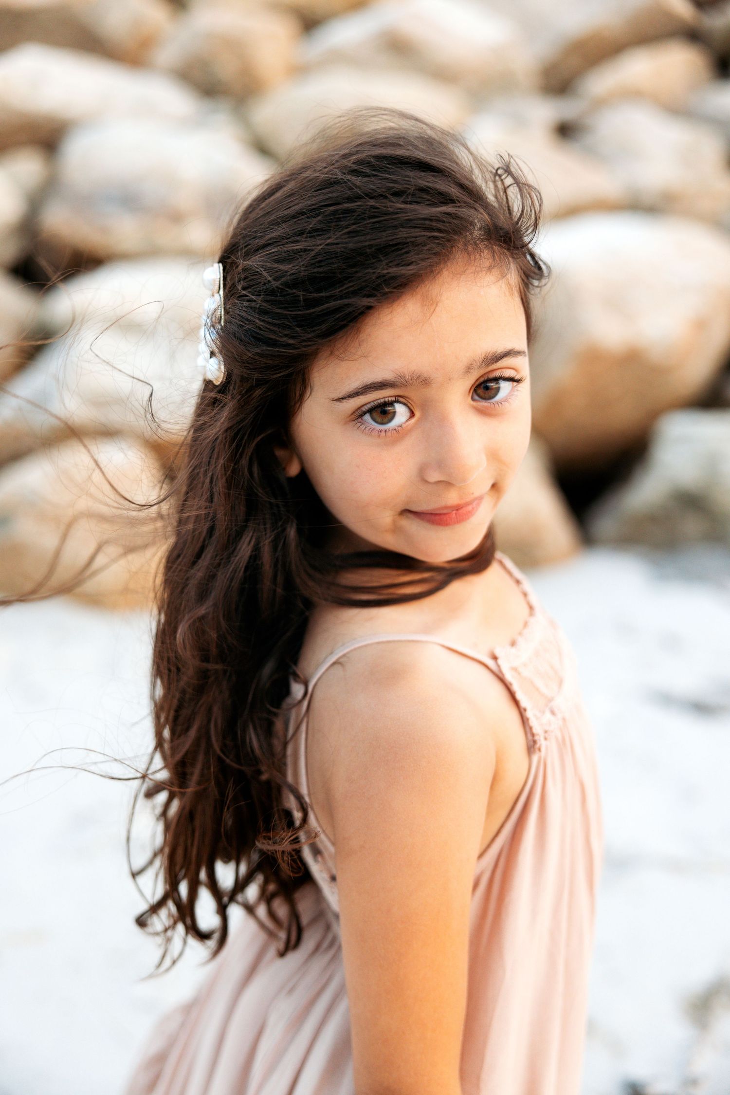 Child in a pale pink dress poses outdoors with wind-blown dark hair against a soft blurred background.