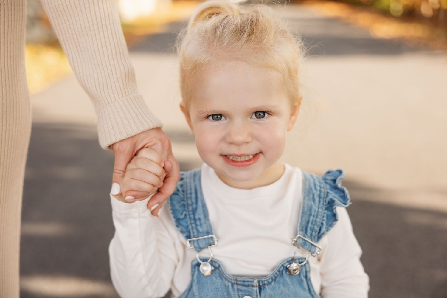 Toddler in denim overalls and cream sweater walking outdoors in autumn setting.