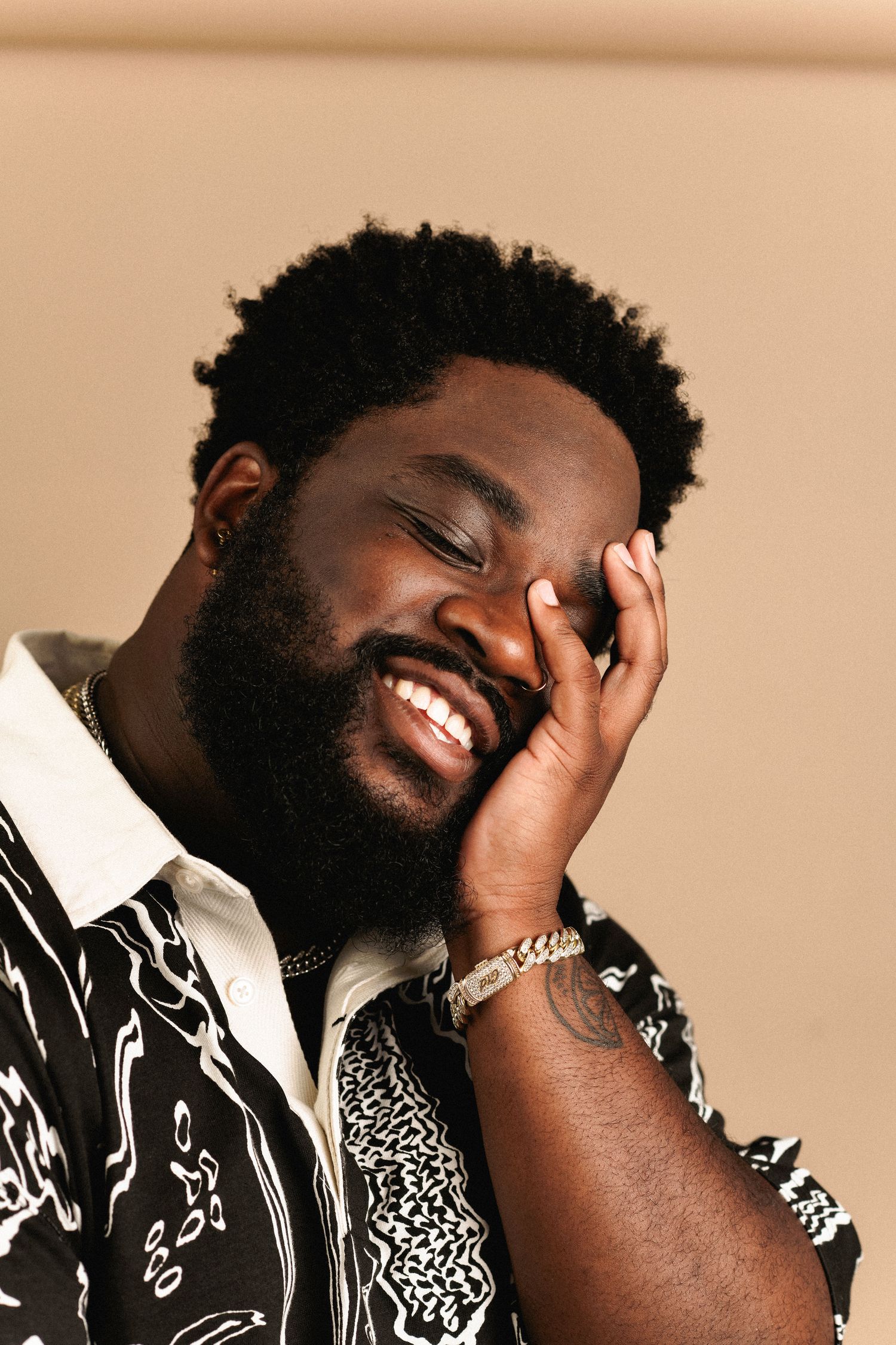 Smiling bearded Black man in patterned shirt resting hand on face with gold jewelry.