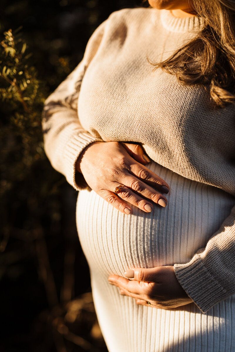 Close-up of hands cradling a pregnant belly in a cream ribbed sweater dress.