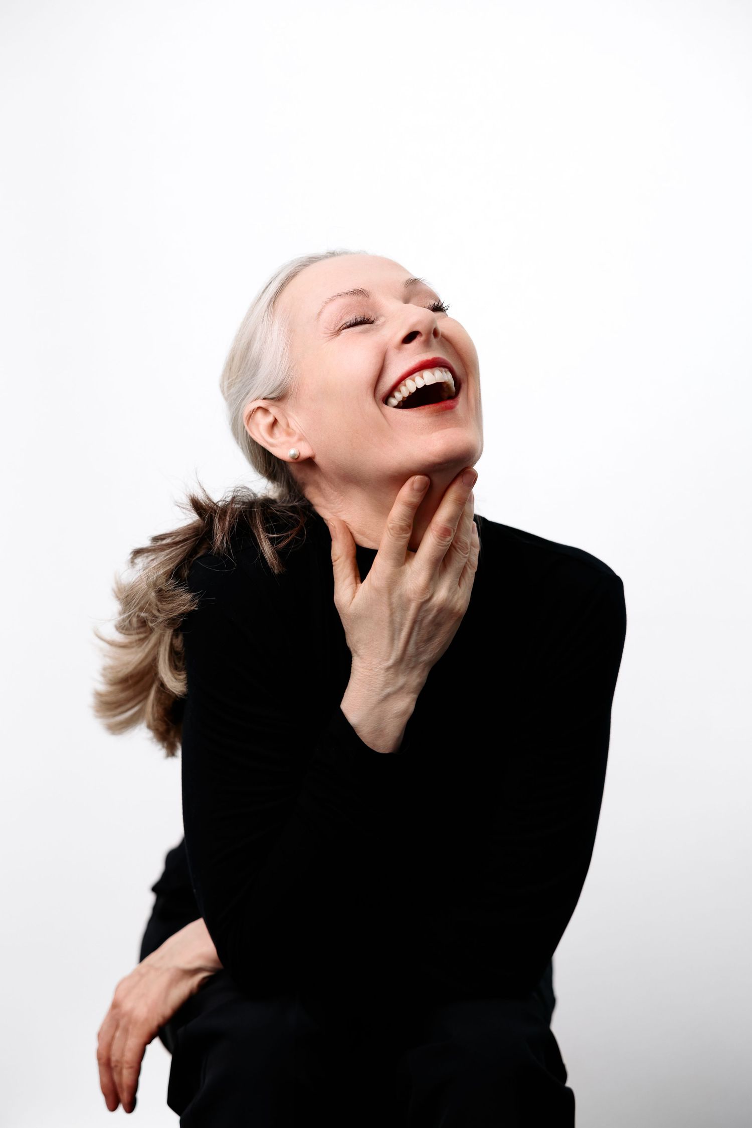 An older woman with gray hair laughs joyfully, wearing a black turtleneck, hand raised to her chin, against a white background.