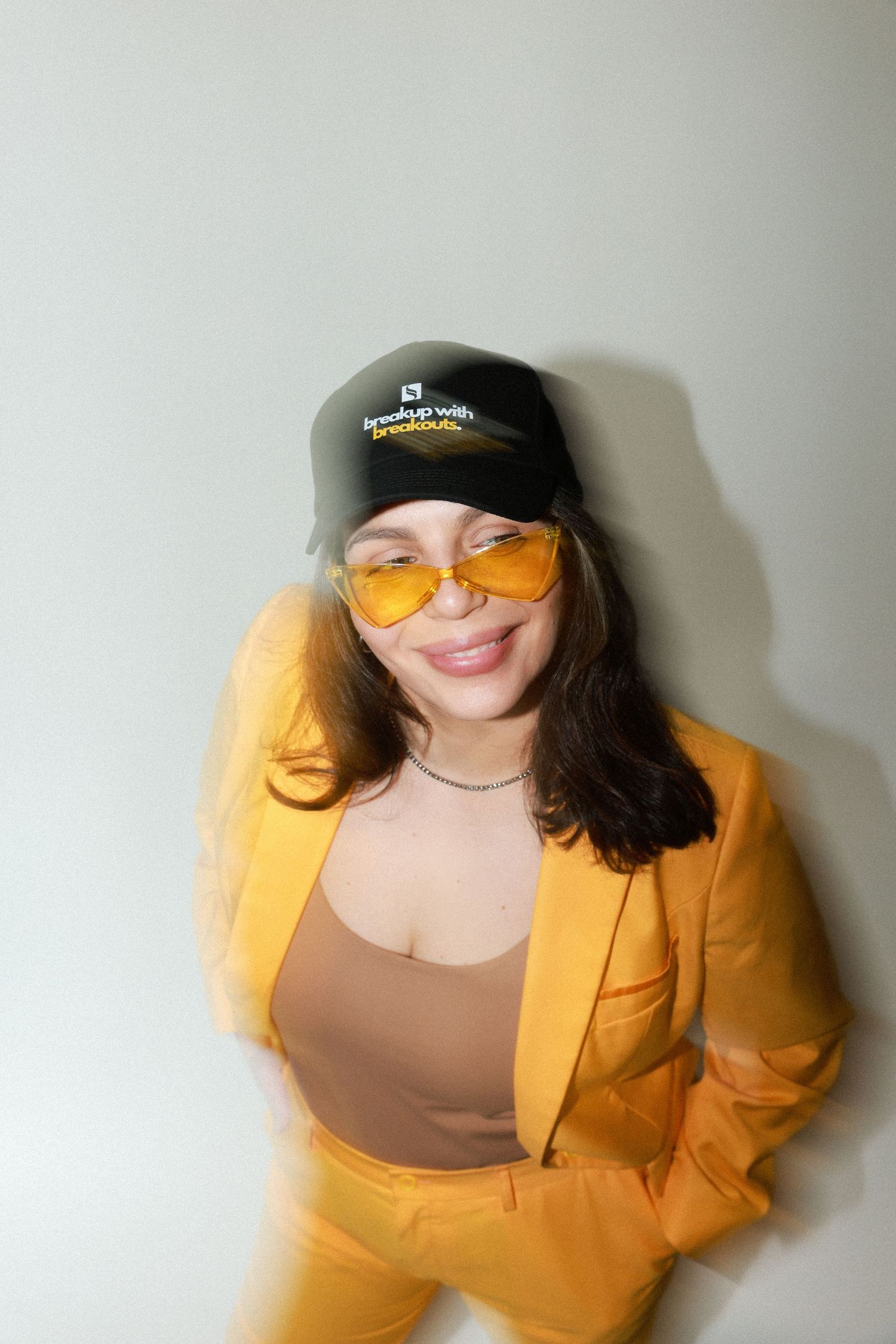 Woman in yellow suit and cap wearing yellow sunglasses smiling against wall.
