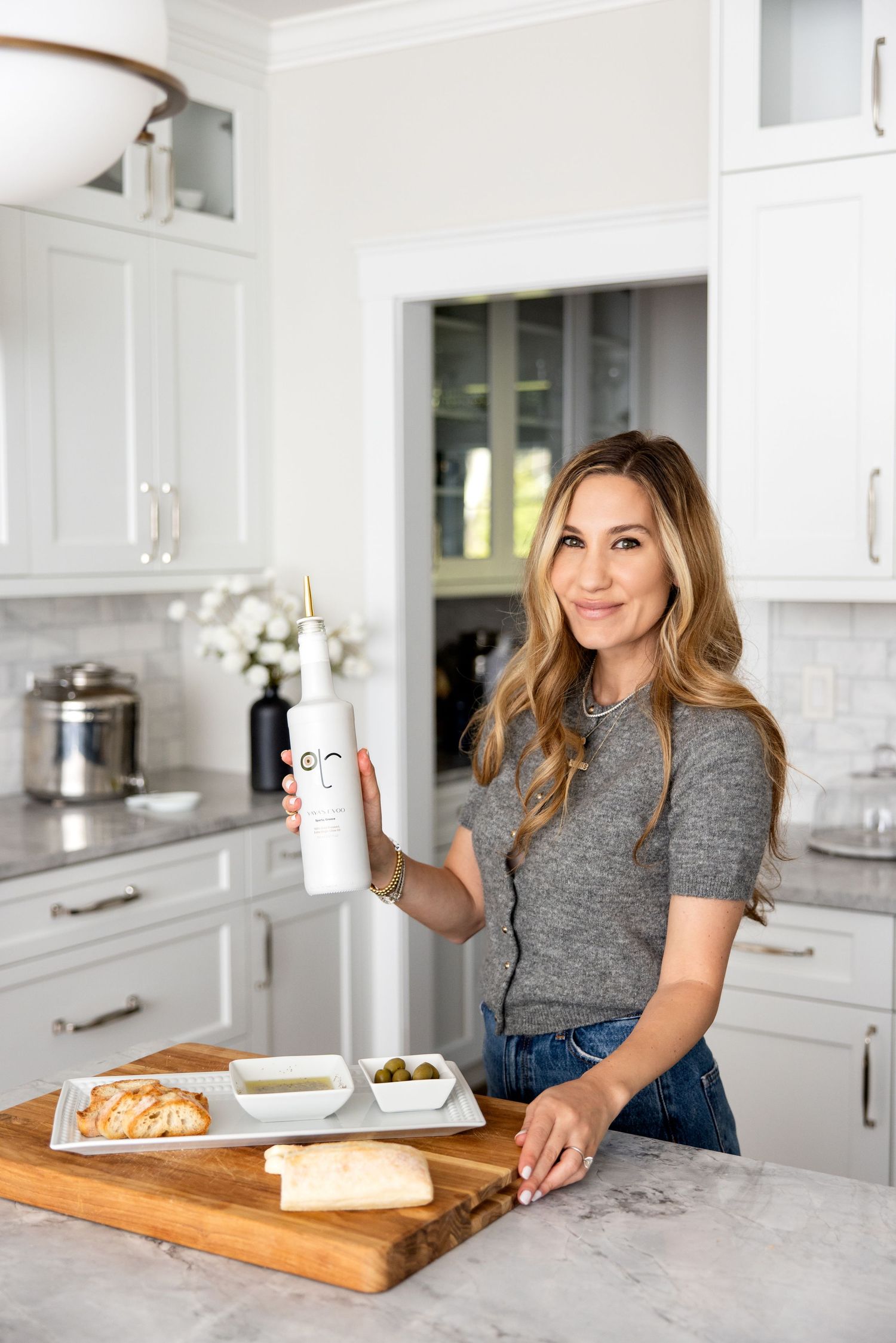 Woman smiling in white kitchen holding branded bottle with charcuterie board on marble counter.