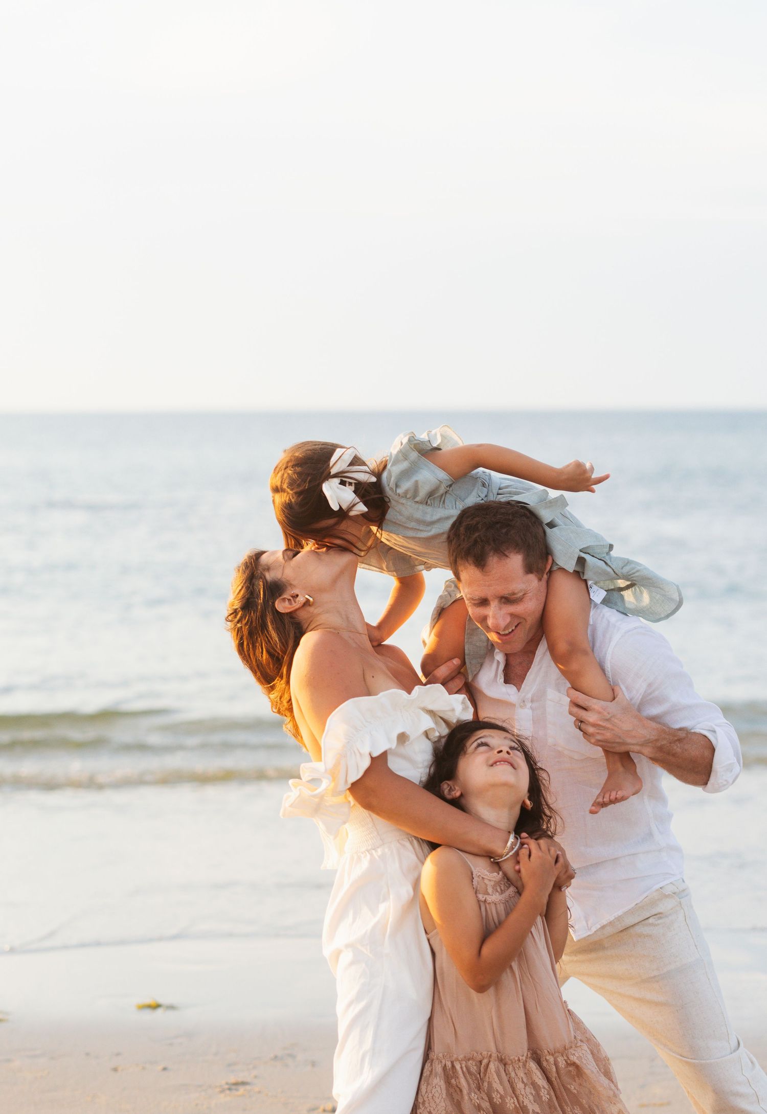 Family dressed in white and beige embraces joyfully while playing on a sunlit beach at sunset.