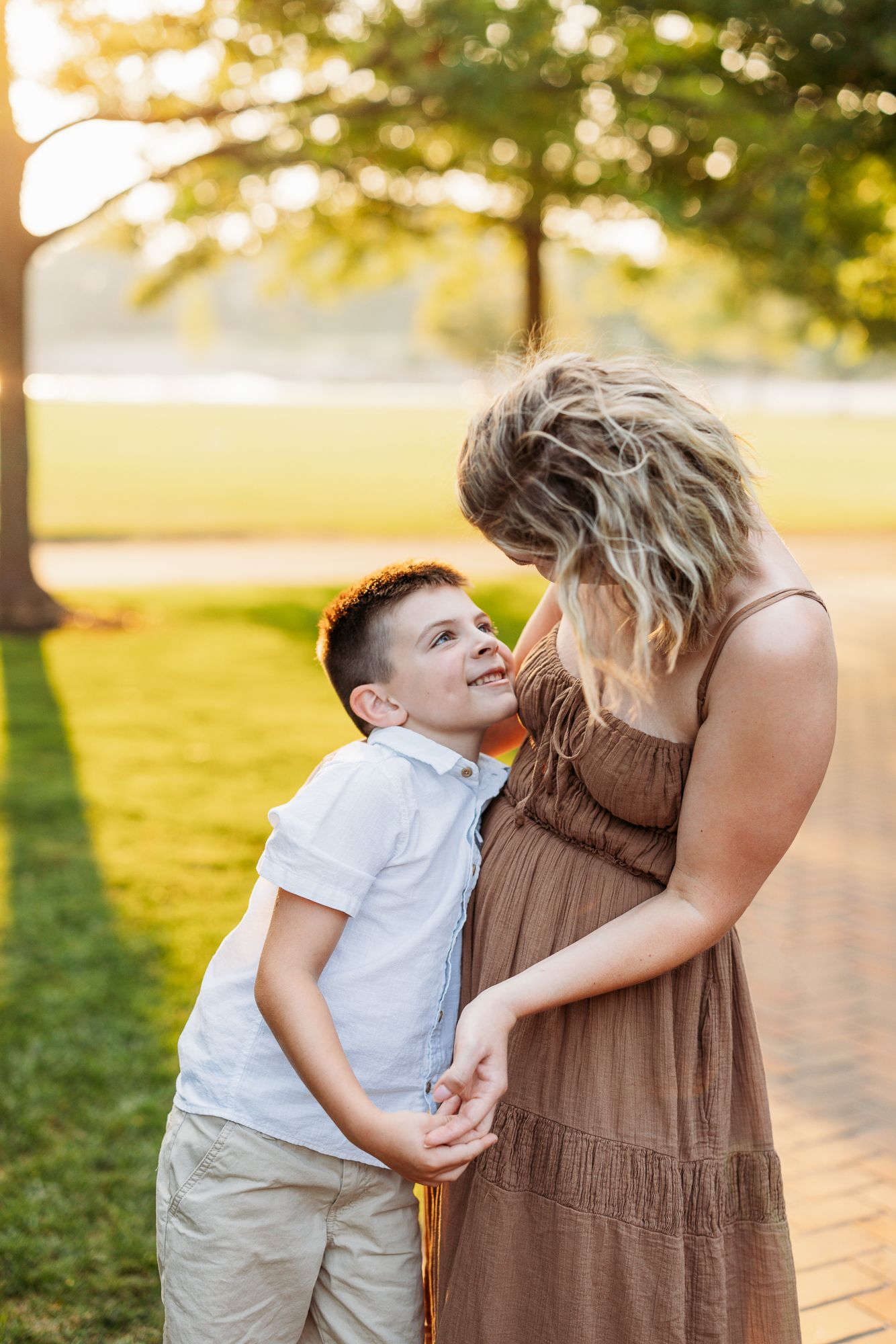 A child in white shirt leans in to give a kiss while standing next to someone in a brown dress at sunset outdoors.
