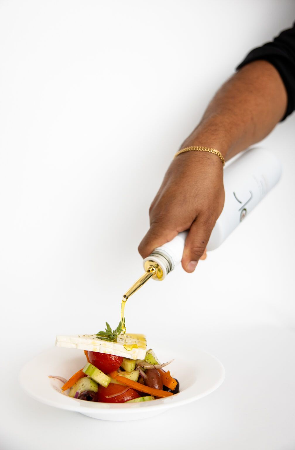 Hand with gold bracelet using utensil to garnish plated food on white surface.
