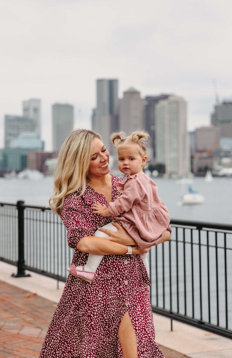Mother and child in matching dresses pose along waterfront with city skyline backdrop.
