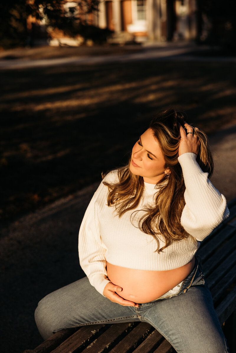 Maternity photo of woman in white sweater relaxing on wooden bench at sunset.