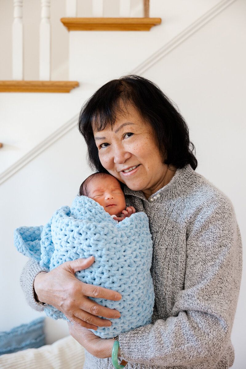 Woman in gray sweater holding sleeping newborn swaddled in blue knit blanket by staircase.