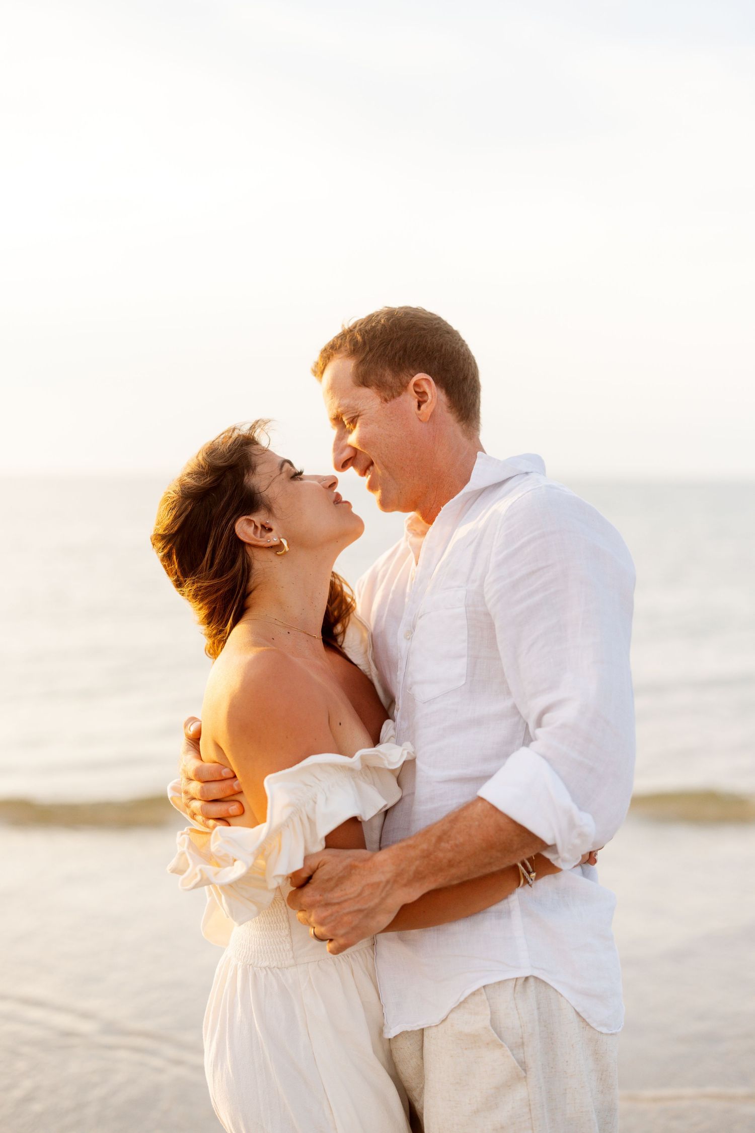 Romantic couple in white outfits embrace and kiss on beach during golden hour sunset.