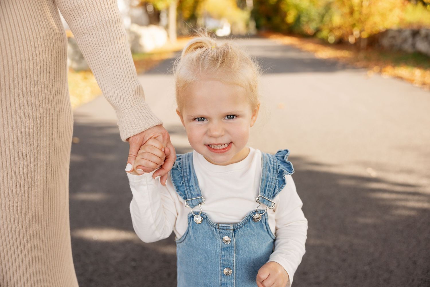 Young child in denim overalls enjoying a walk while holding hands on an autumn path.