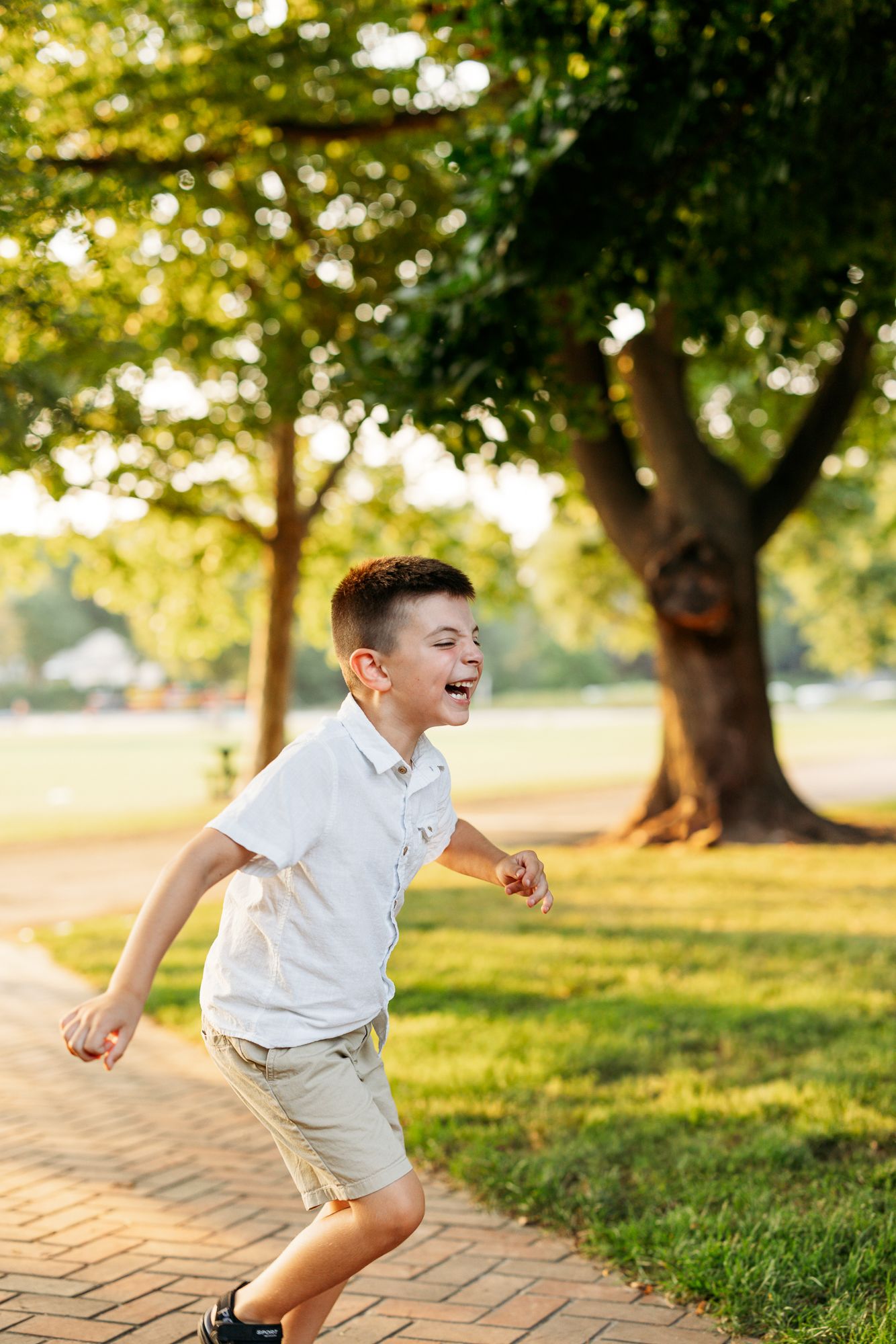 Young child jumps playfully on a brick path in a sunlit park with trees and green grass in the background.