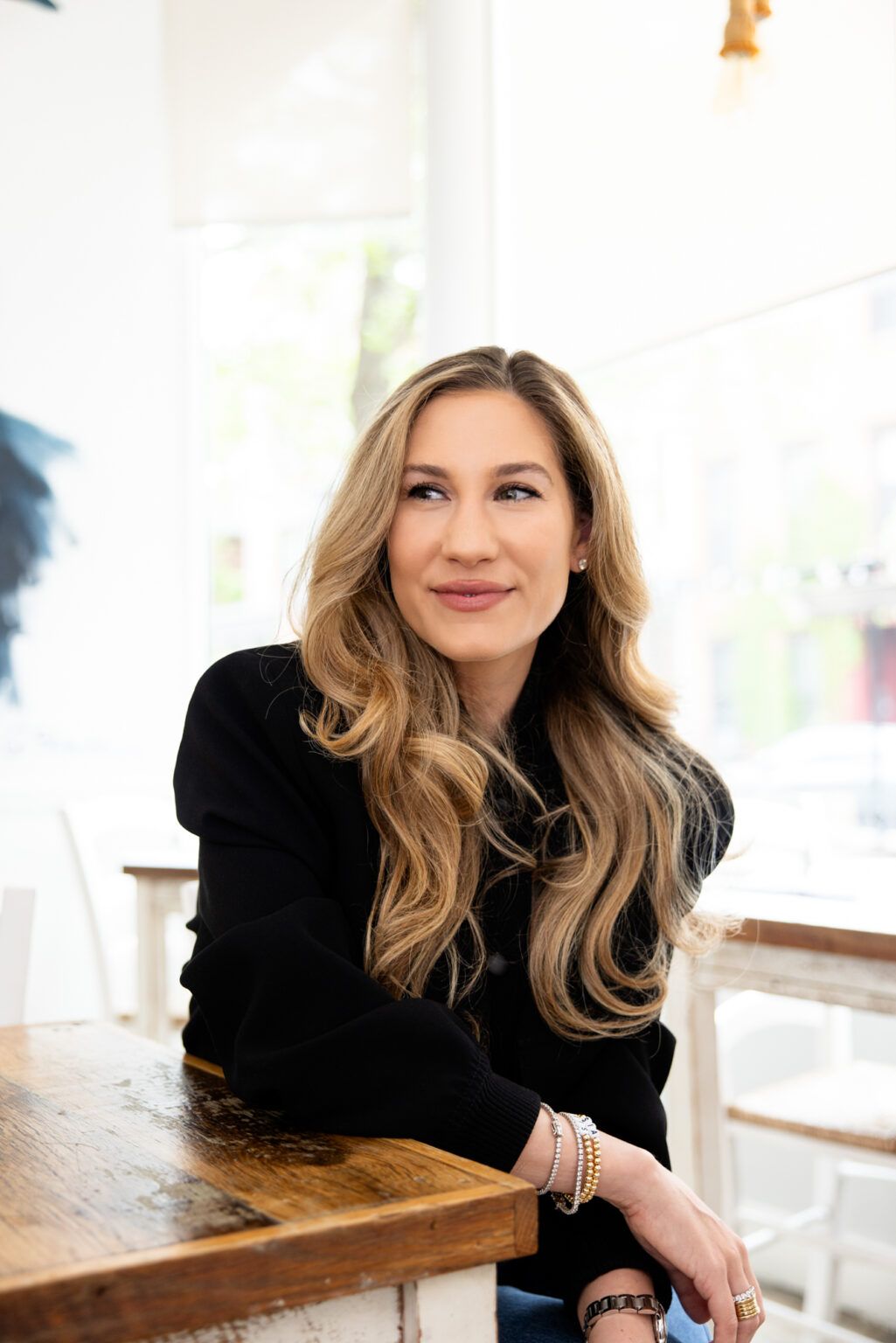 Professional headshot series in bright setting with wavy blonde hair and black top resting near wooden table surface.