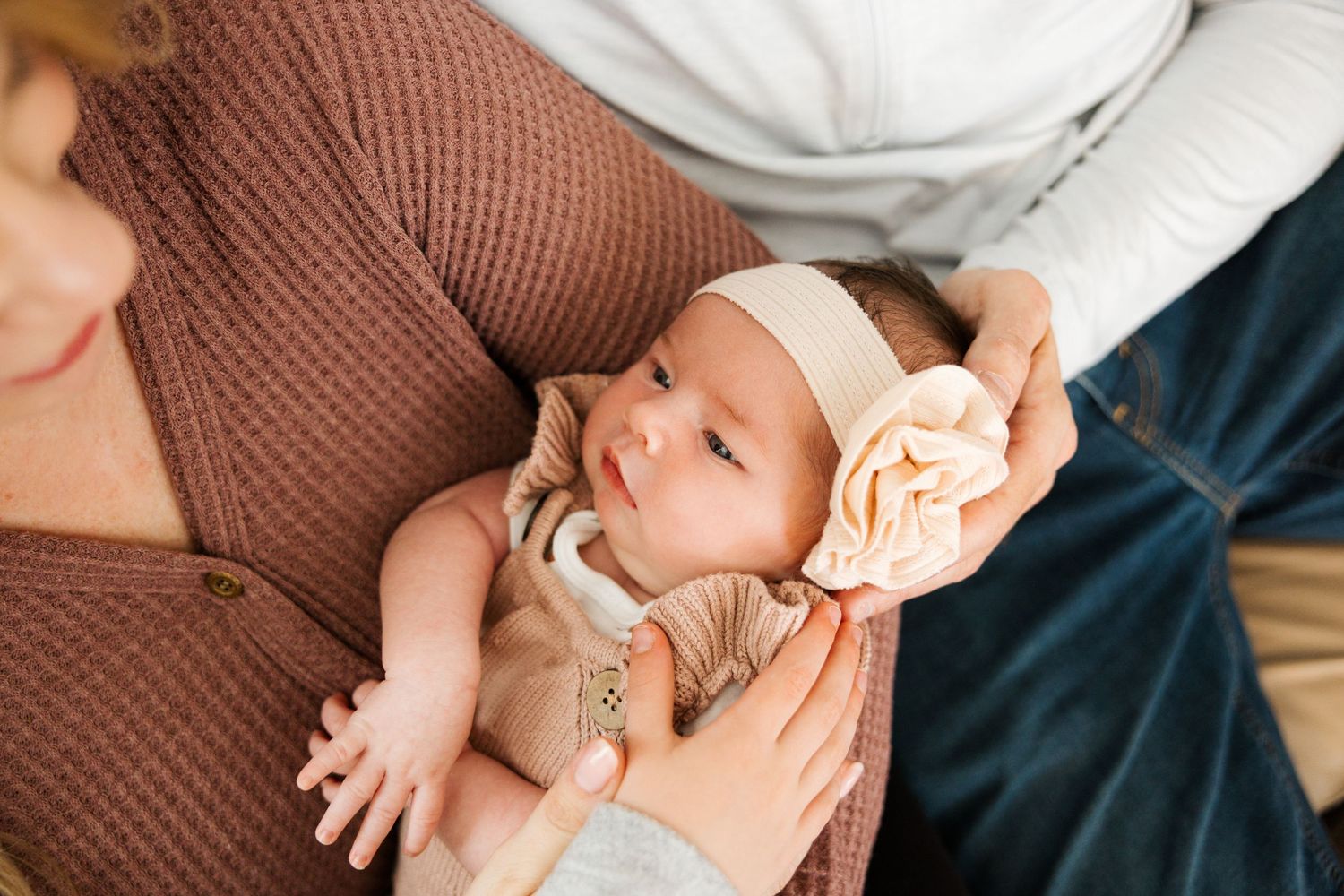 Baby wearing peach floral headband rests peacefully in parent's arms wearing neutral colored clothing.