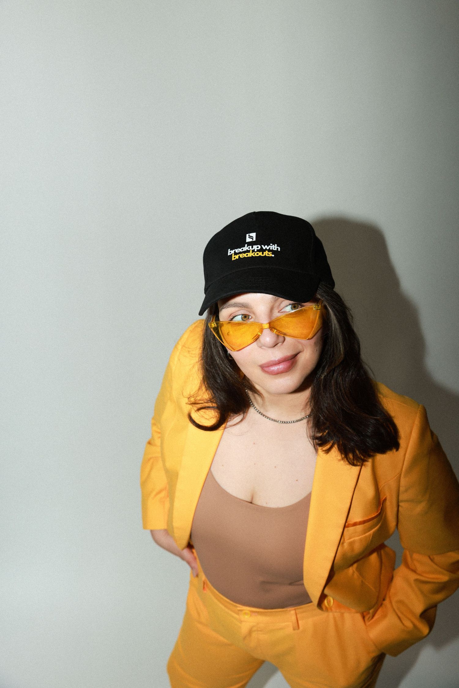 Portrait of person in mustard yellow blazer wearing orange tinted glasses and black baseball cap against white wall.