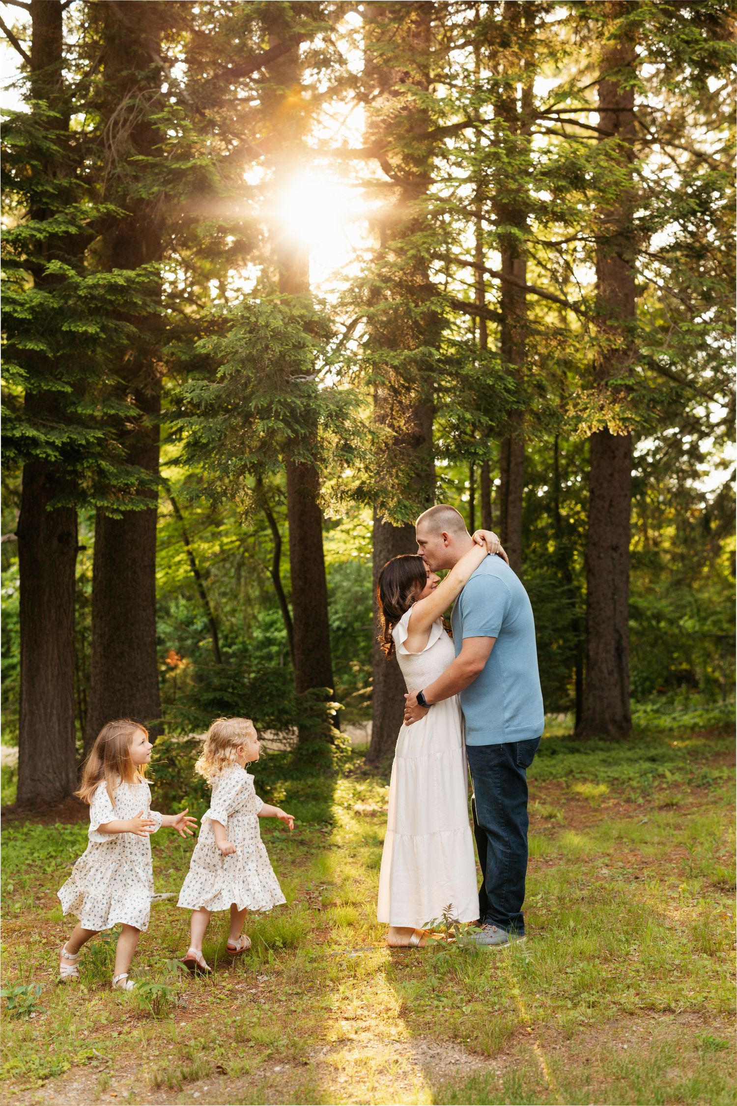 Family with two young girls dancing in sunlit forest clearing, parents embracing at golden hour.