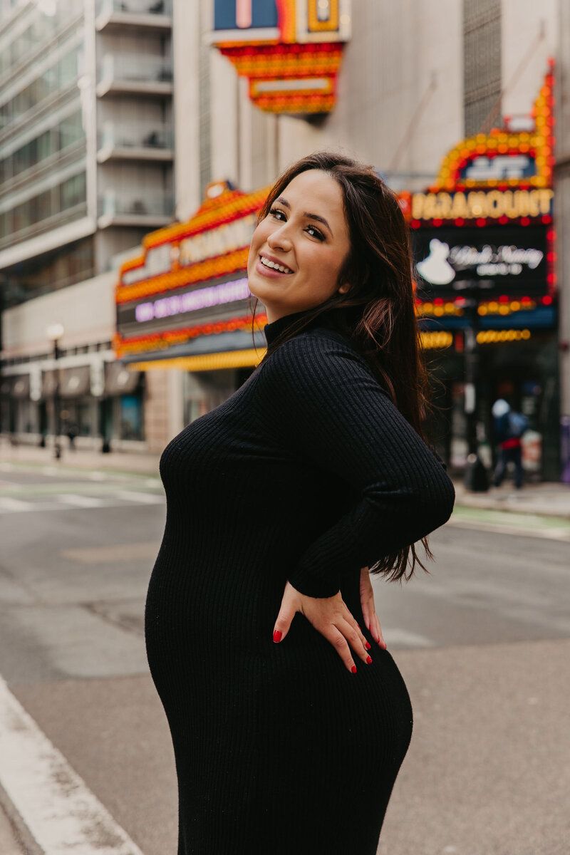 Woman in black sweater posing playfully on urban street with neon signs.