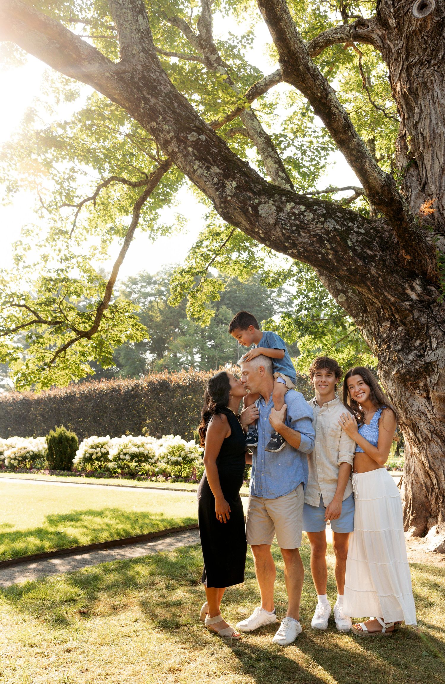 A family group poses playfully together under large trees in a sunny outdoor setting with green grass.