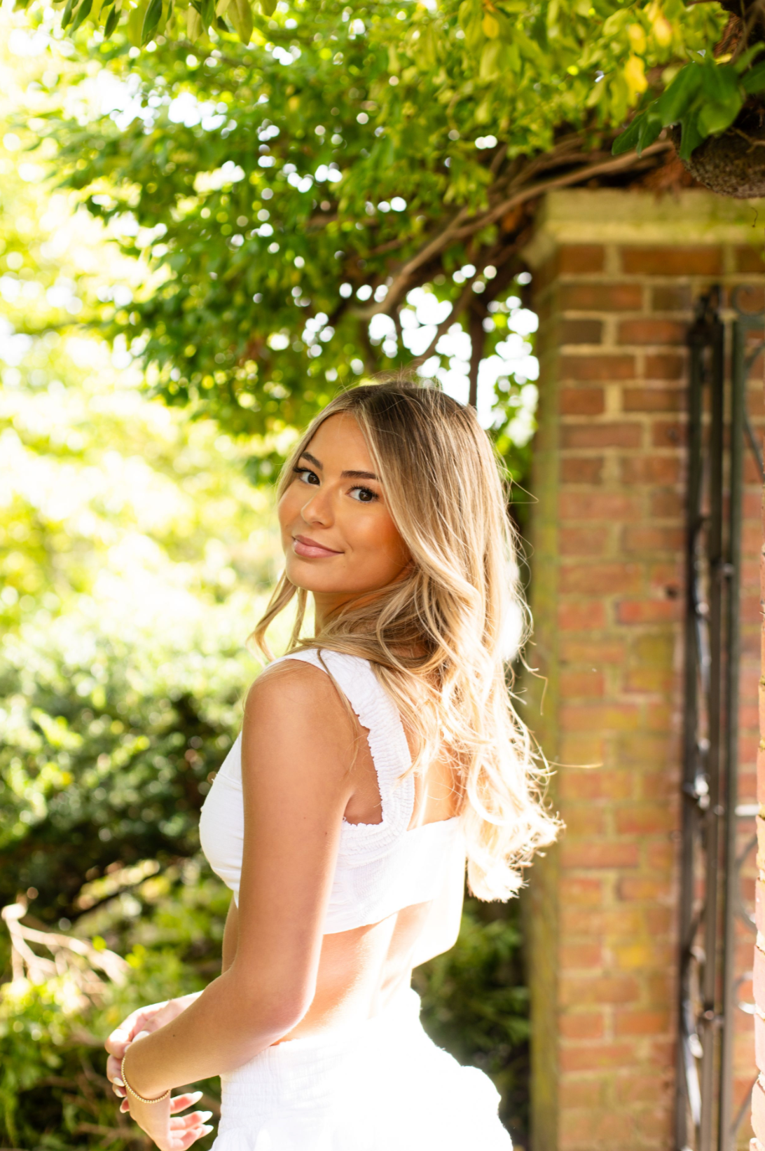 Young woman with blonde hair in white dress poses outdoors near brick wall with lush green foliage.