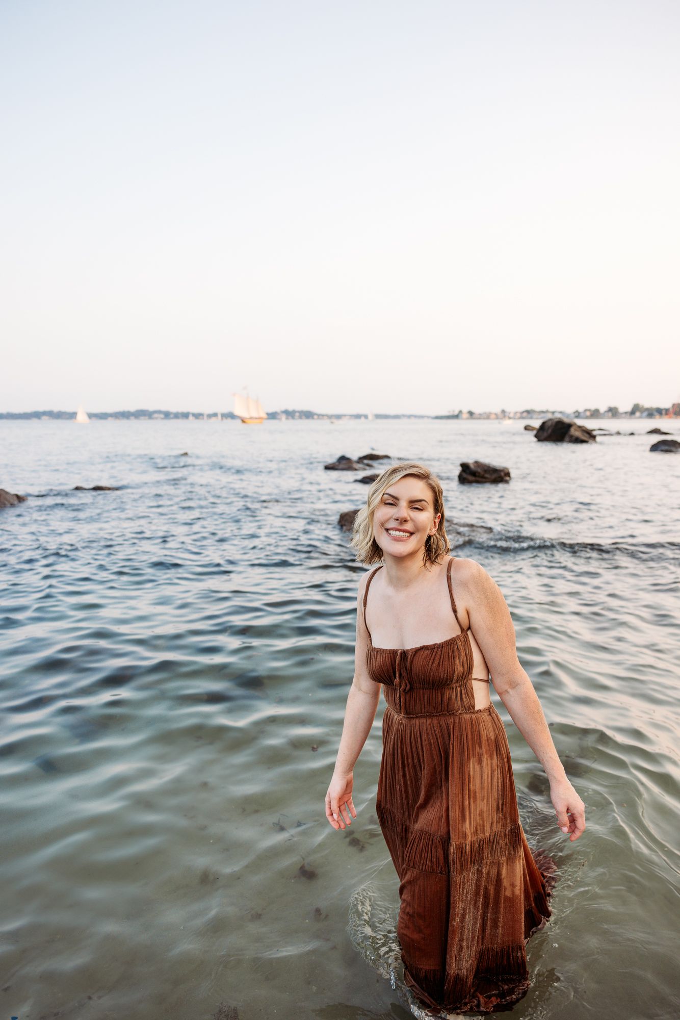 Person in brown dress splashing and laughing in ocean water during sunset.