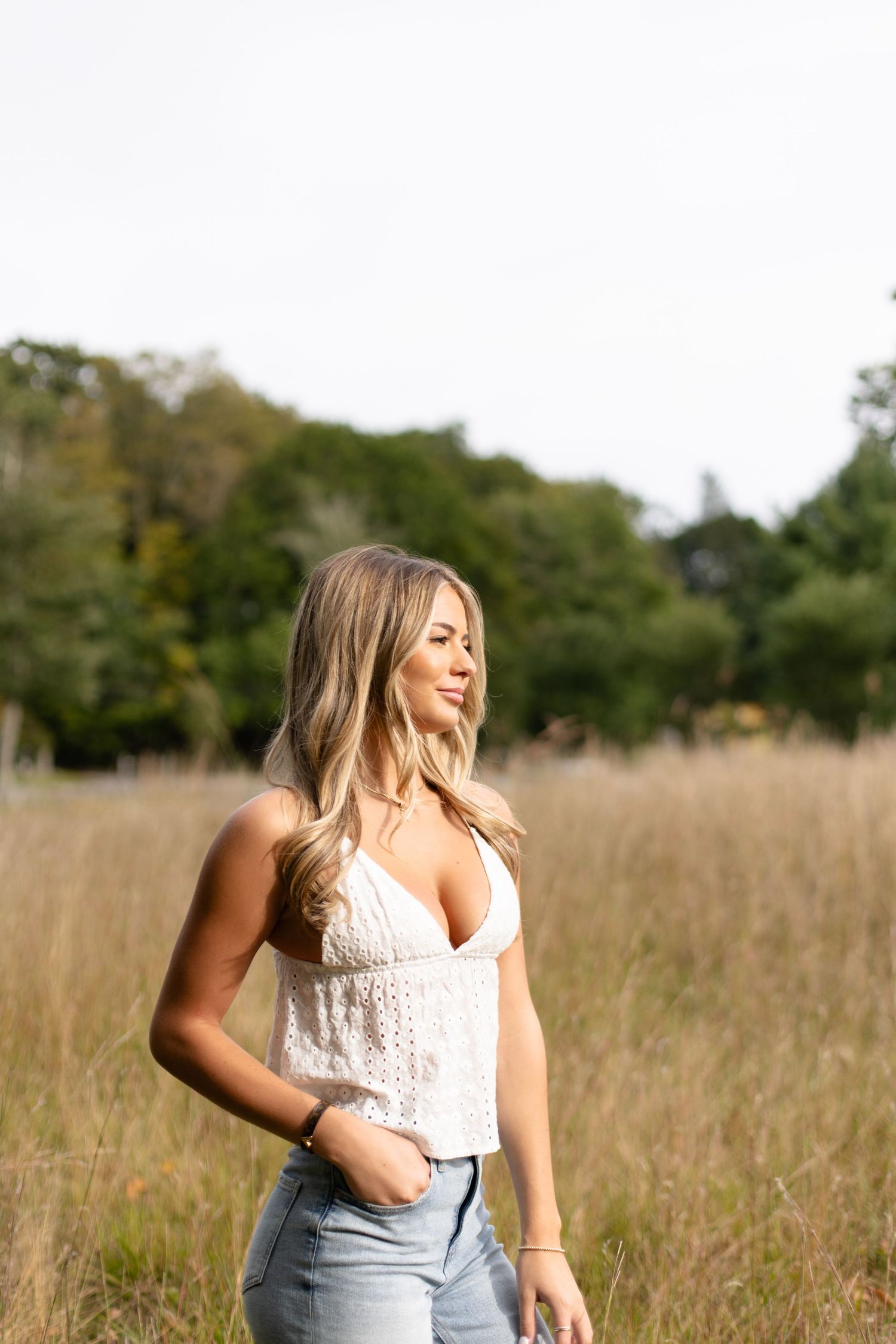 A person wearing a white lace top and jeans stands smiling in a sun-lit field of tall grass.