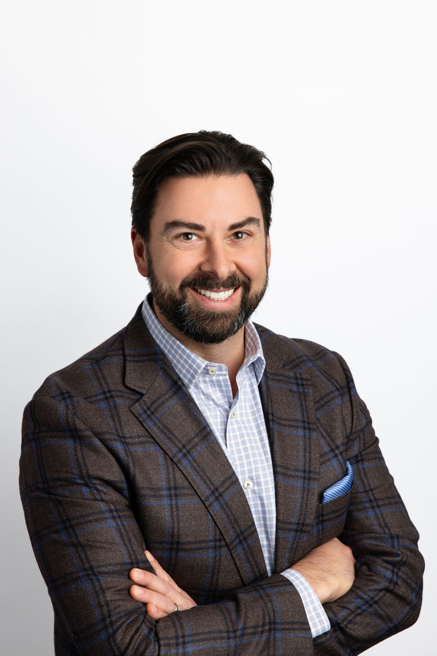 Smiling bearded man in plaid blazer with arms crossed, professional headshot on white background.