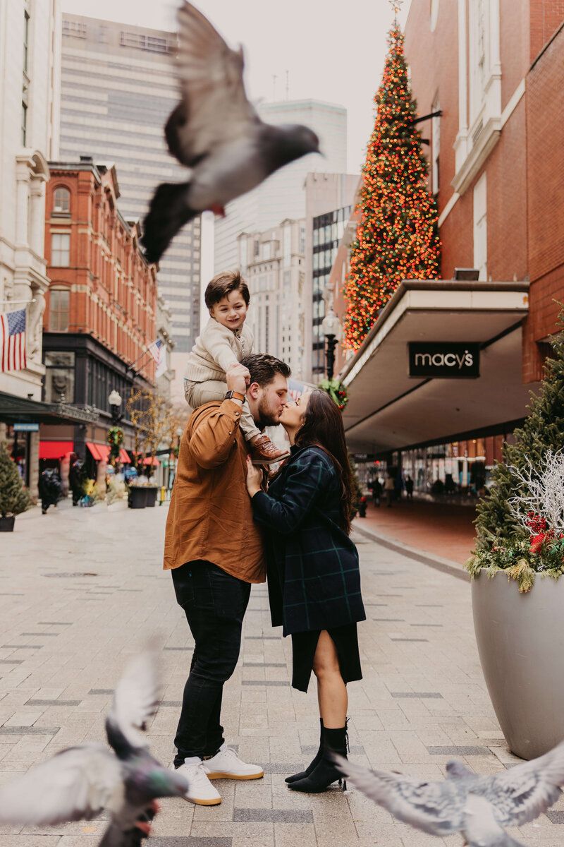 A couple kisses on a city street near Macy's, man holding toddler, pigeons flying, Christmas tree in background.