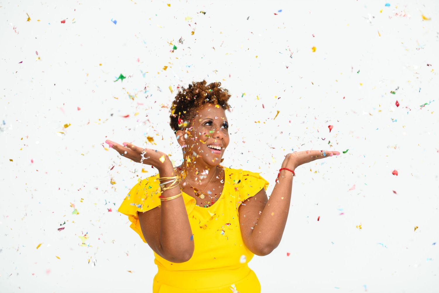 A joyful woman in a yellow dress celebrates with colorful confetti falling around her on a white background.