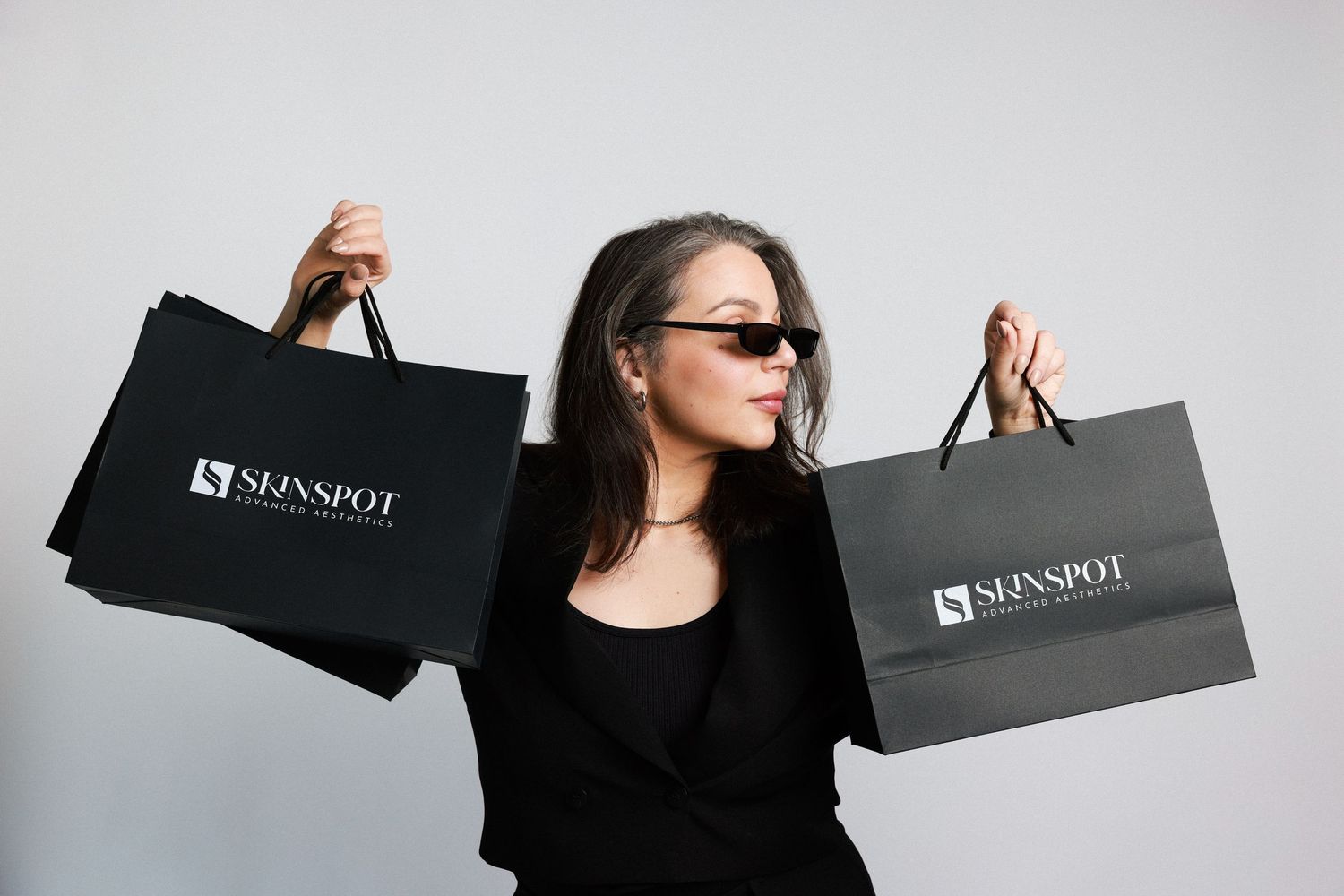 Woman in sunglasses holding two black Skinspot shopping bags against gray background.