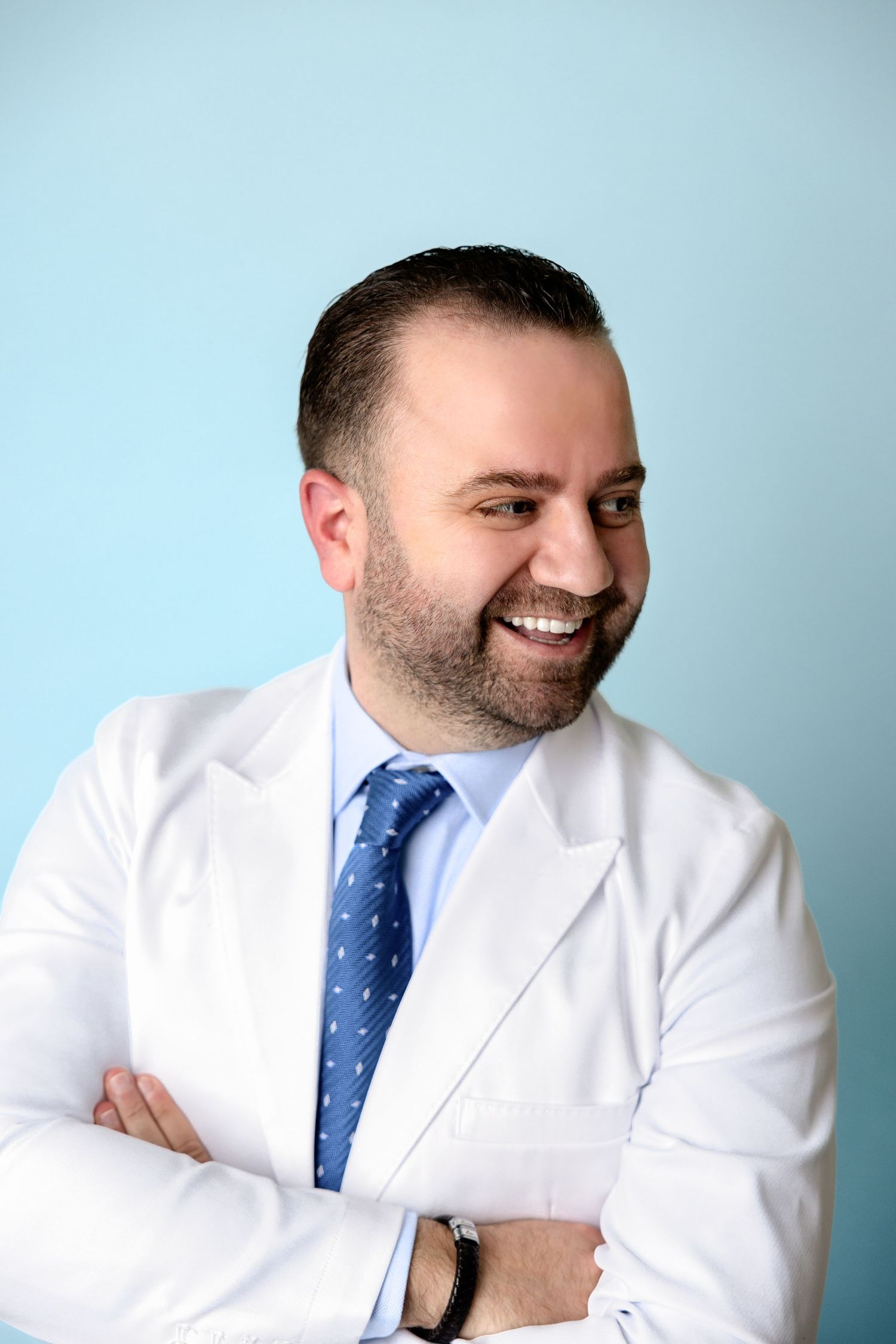Smiling male doctor in white lab coat and blue tie with arms crossed against light blue background.